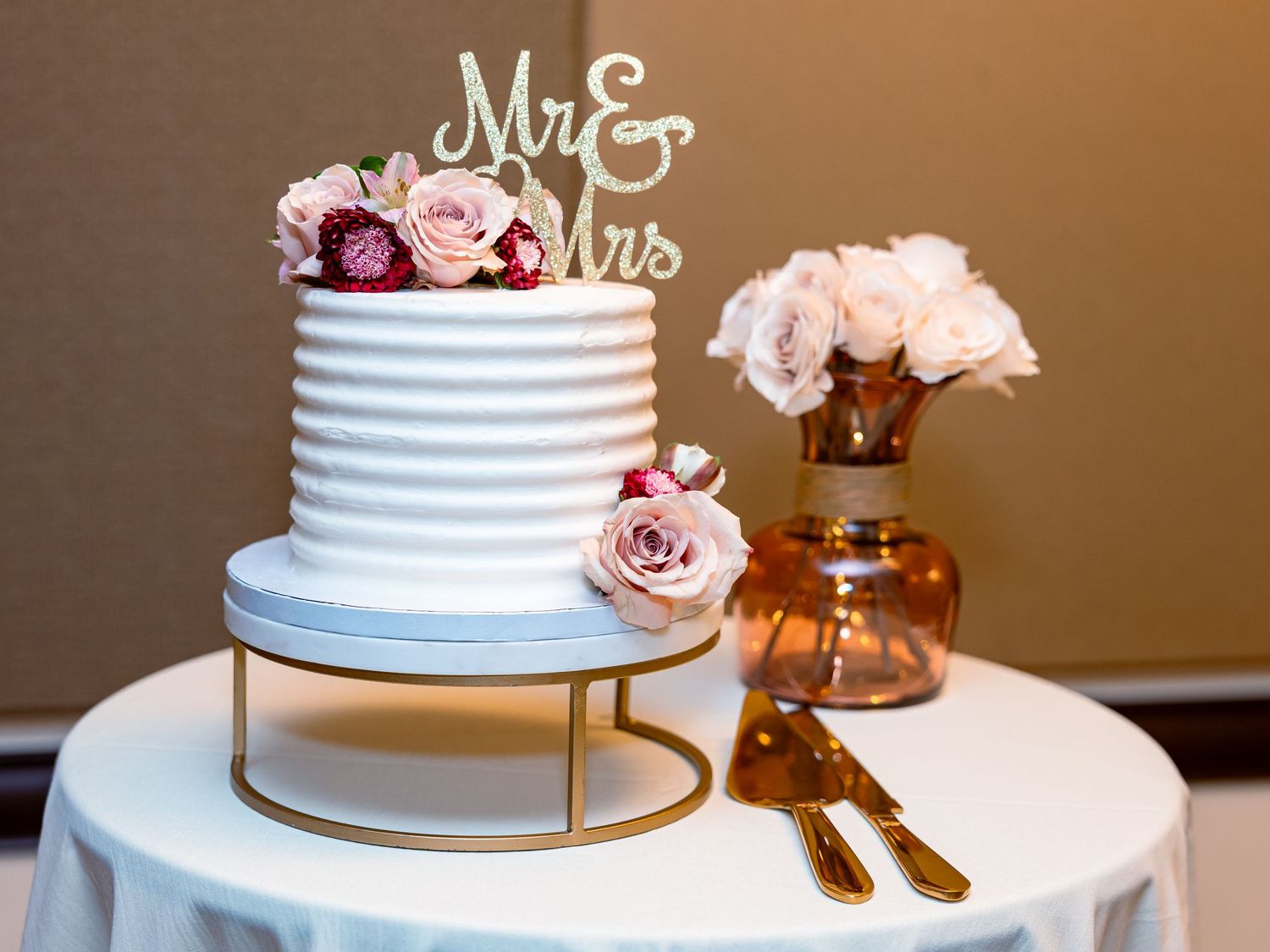 Elegant white wedding cake with pink roses on gold stand next to amber vase with matching floral arrangement.