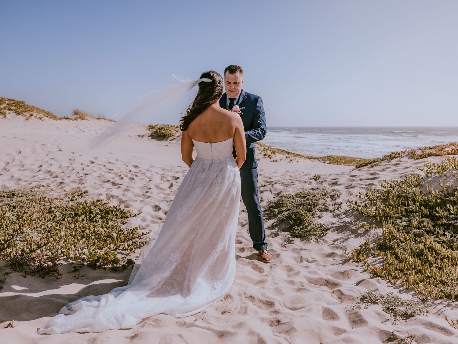 Couple in wedding attire share an intimate moment on a sandy beach with ocean waves in the background.
