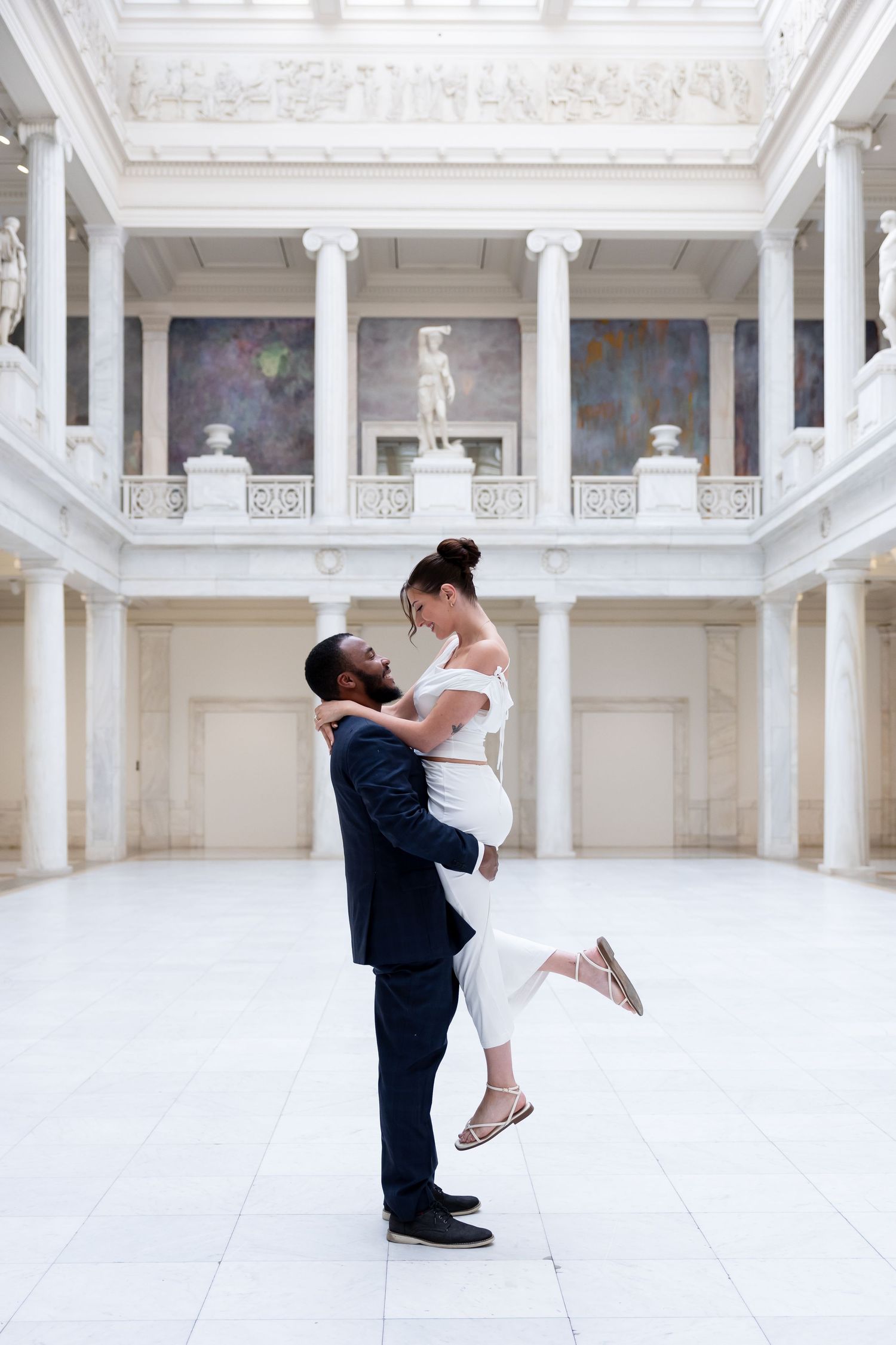 Groom lifts bride in white outfit for a romantic kiss inside an elegant white marble hall with columns.