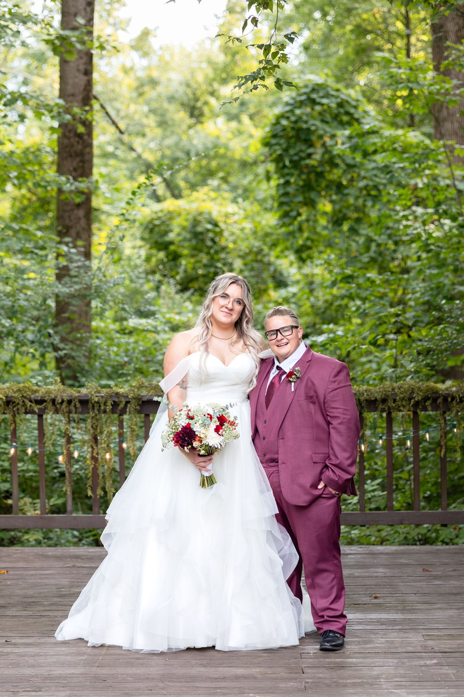 Two brides pose on a woodland deck, one in a white gown holding a bouquet, the other in a burgundy suit.