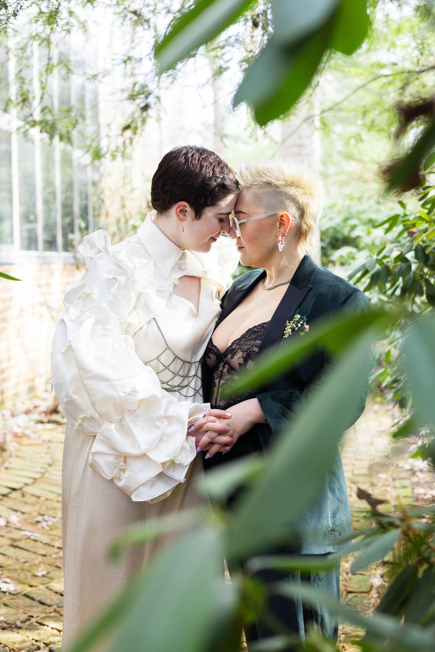 Two brides in unique wedding attire share intimate moment surrounded by lush green foliage in greenhouse.