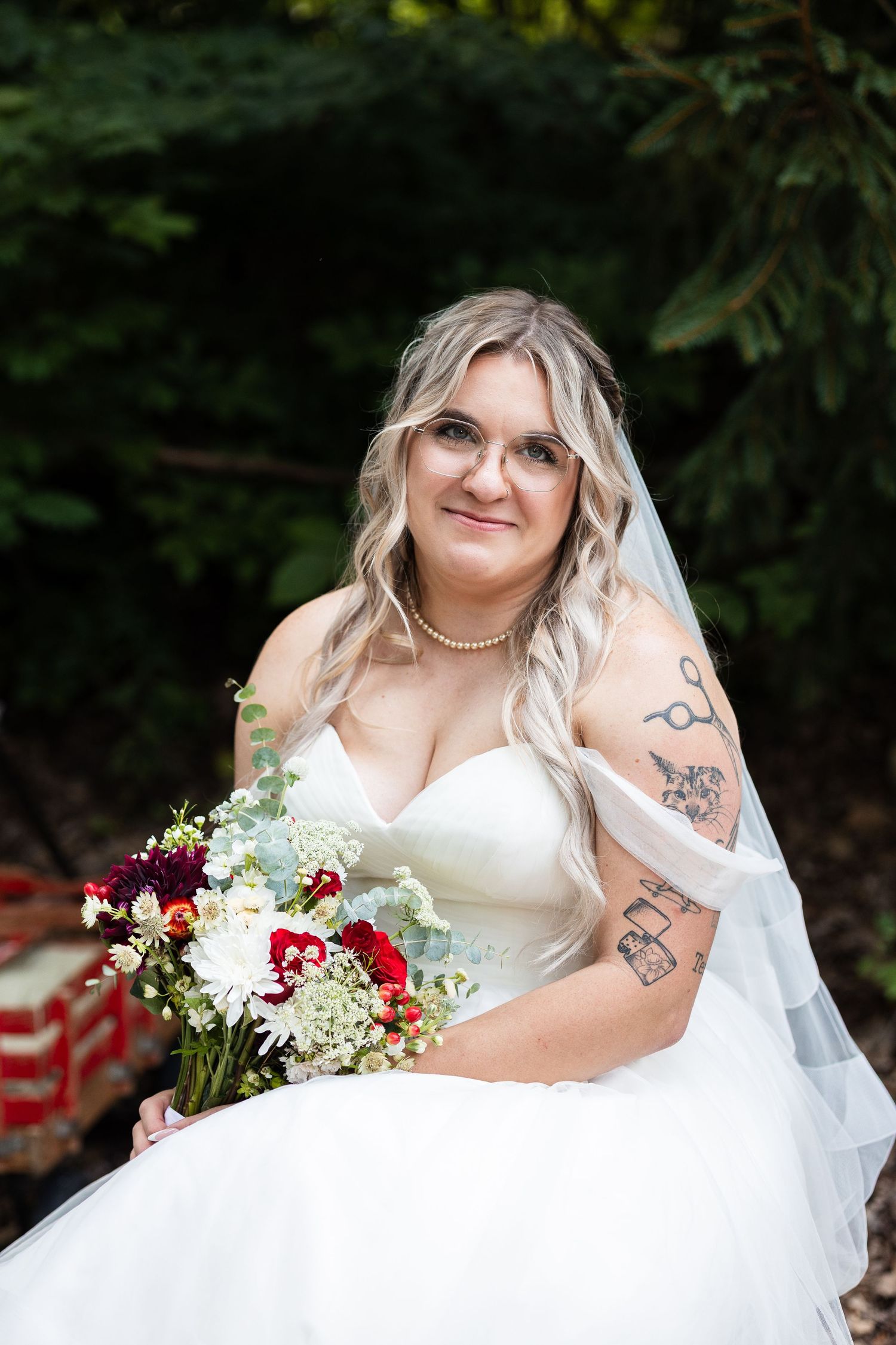 Tattooed bride in strapless white gown with veil and pearl necklace holding colorful bouquet.