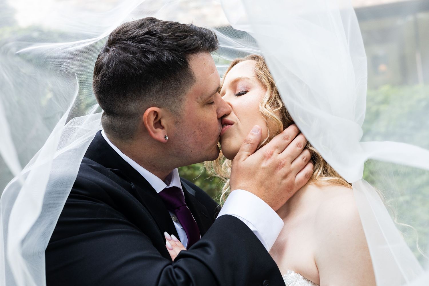 Bride and groom sharing romantic kiss under flowing white veil on wedding day.