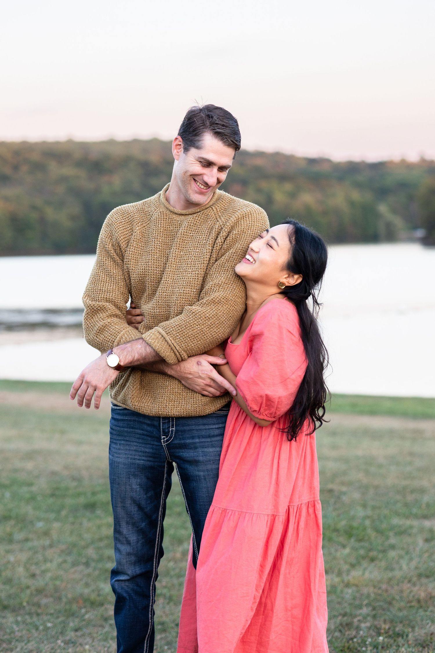 Happy interracial couple embraces outdoors near a lake at sunset, woman in coral dress laughing joyfully.