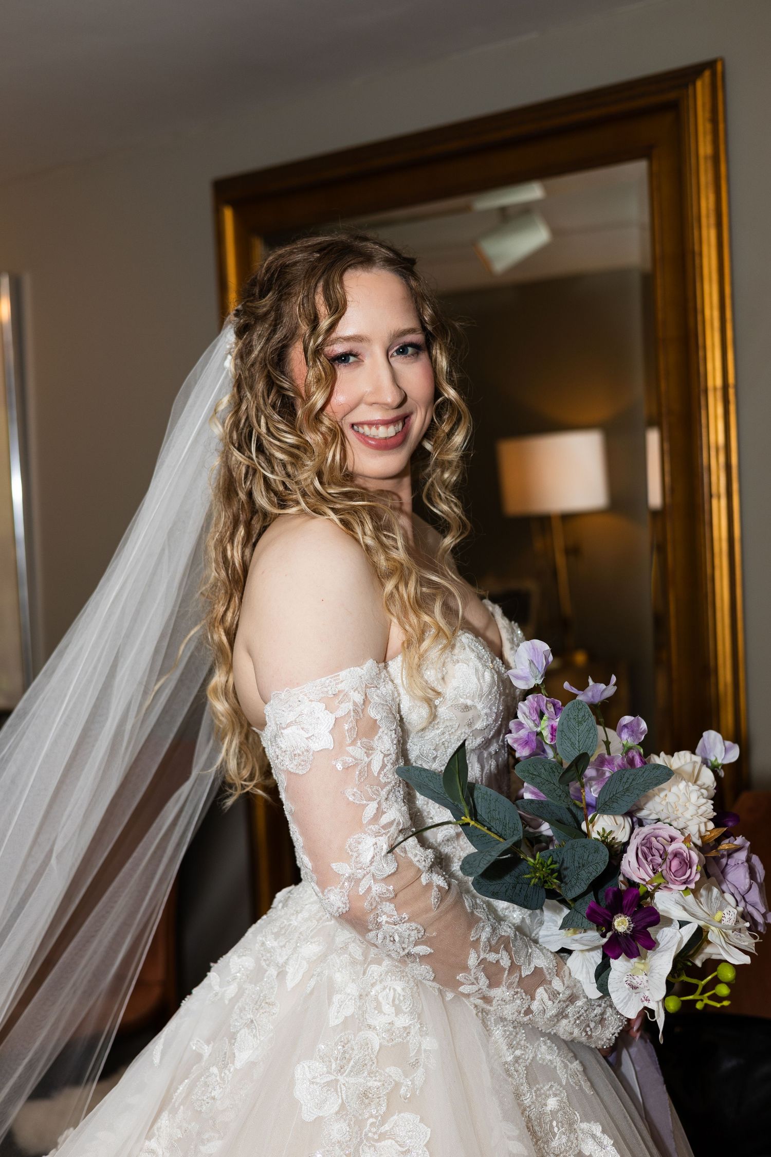 Radiant bride in lace off-shoulder gown with veil holding purple and white bouquet indoors.
