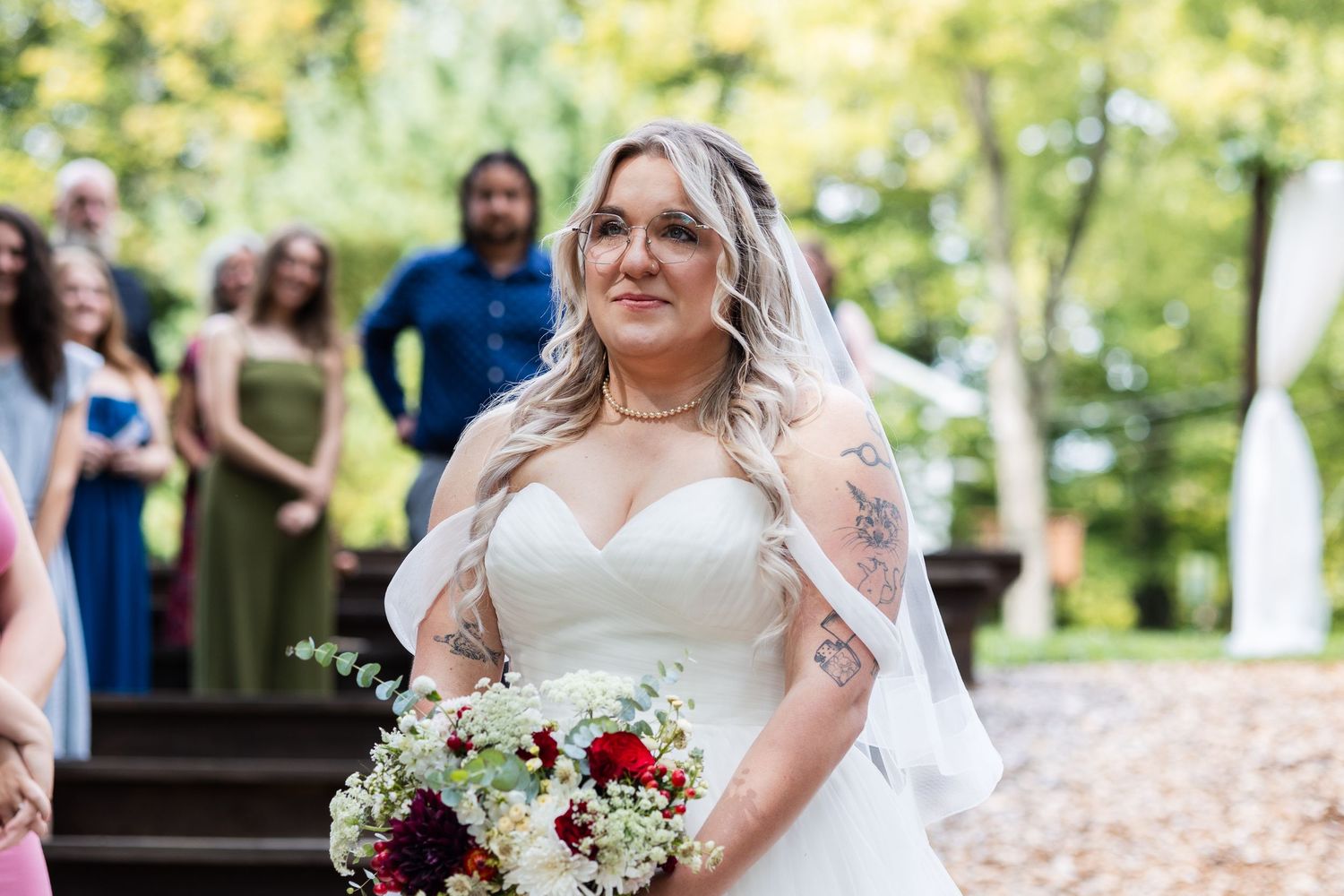 Tattooed bride in strapless gown with veil holding red bouquet walking down aisle.