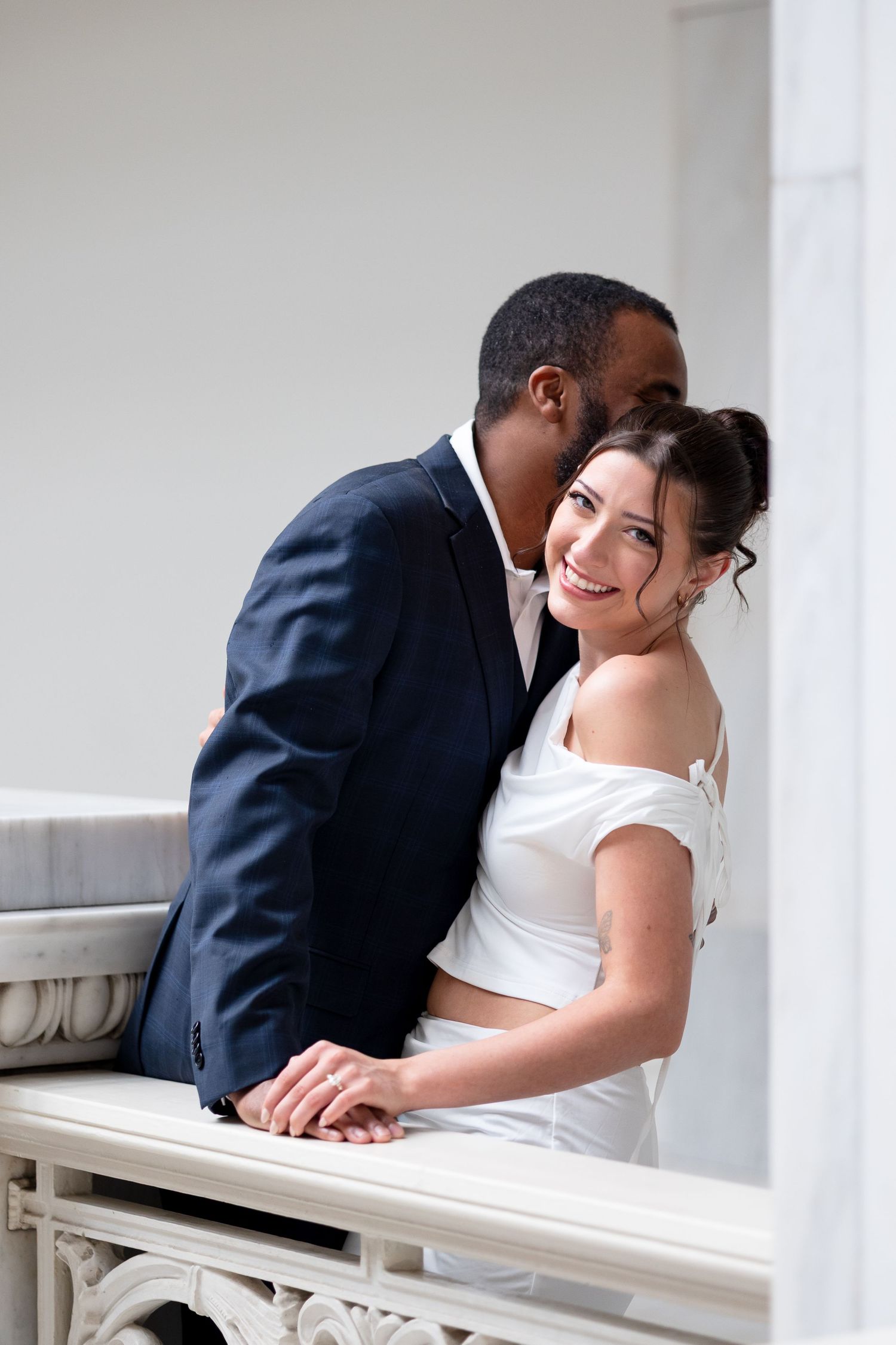 Interracial couple shares a tender moment on a marble balcony, groom kissing smiling bride on the cheek.