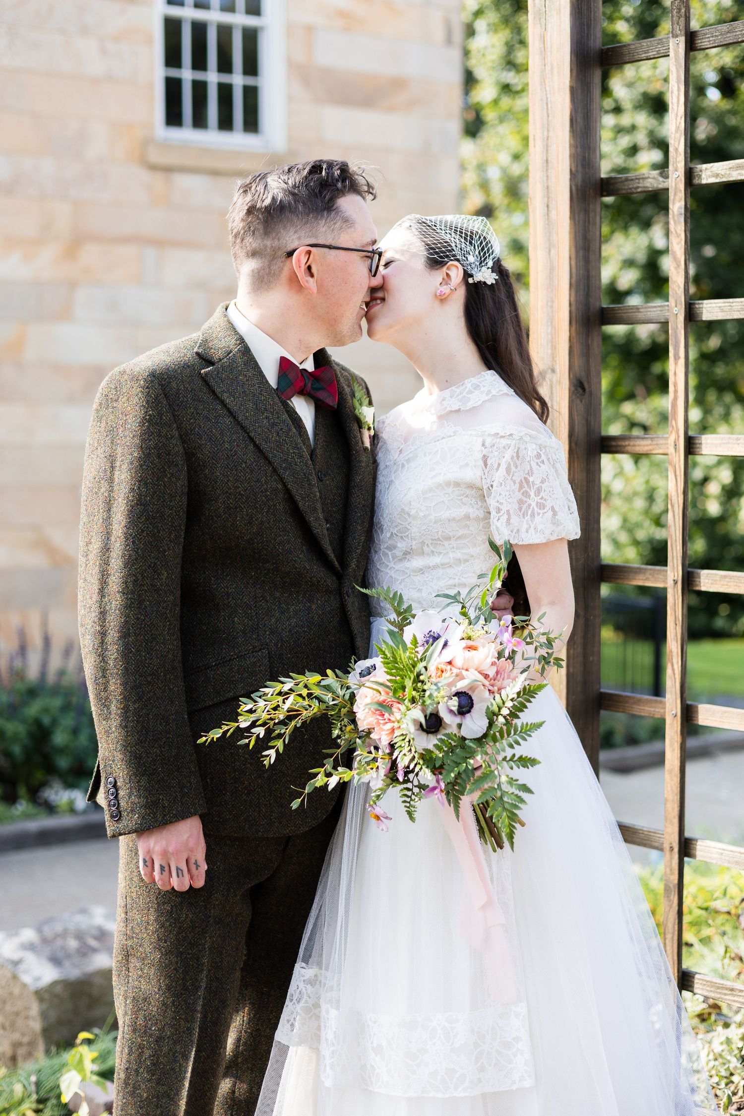Couple sharing tender kiss outdoors, groom in tweed suit, bride holding colorful bouquet.