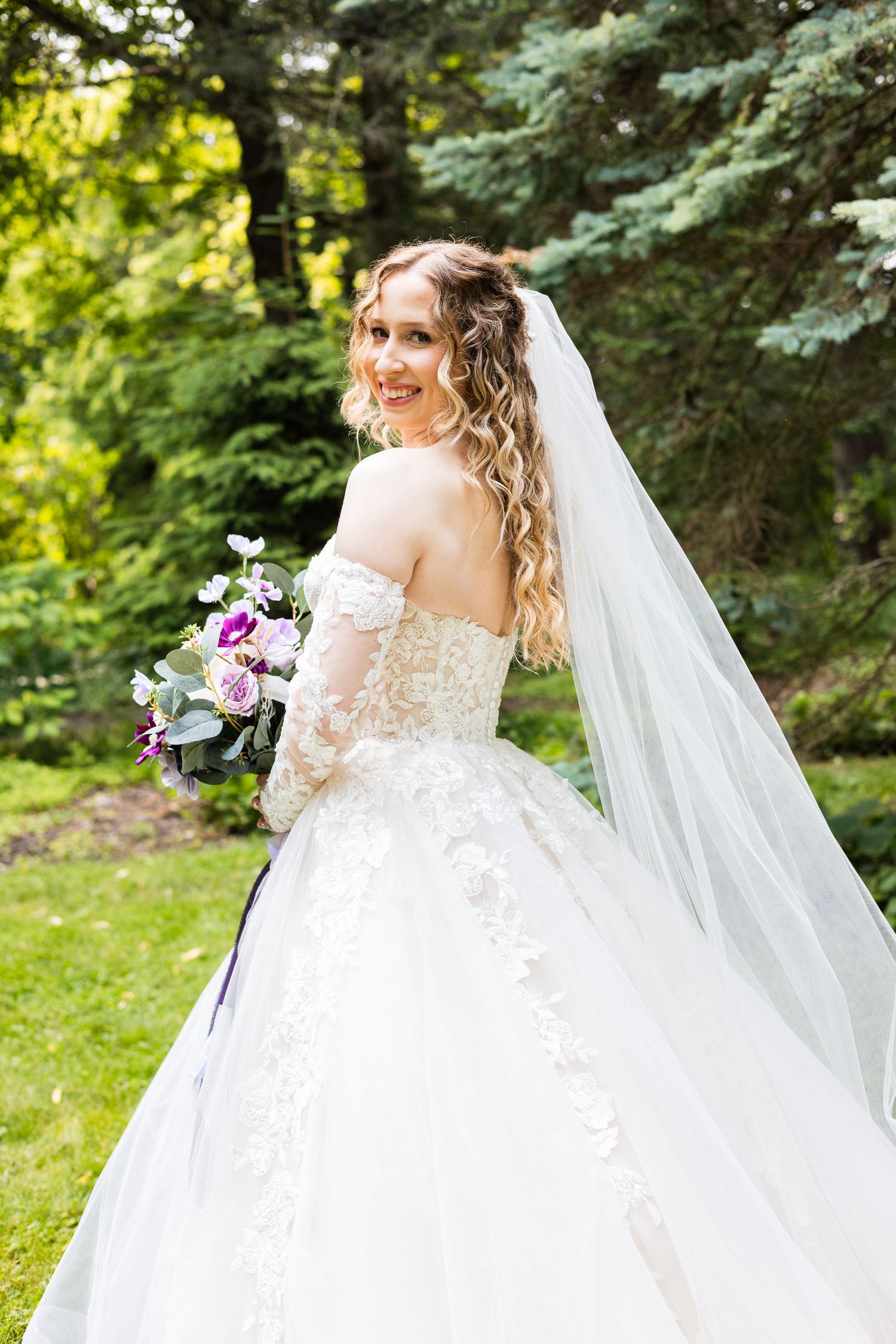 Smiling bride in off-shoulder lace ball gown with veil holding purple bouquet outdoors.