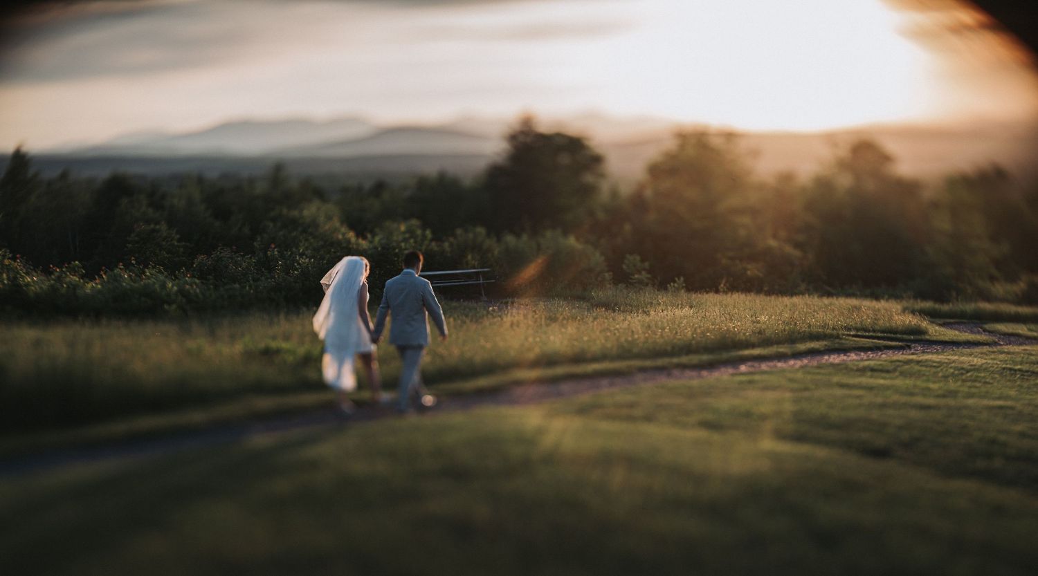 Couple walking hand in hand on a winding path through green fields at golden sunset with mountain views.