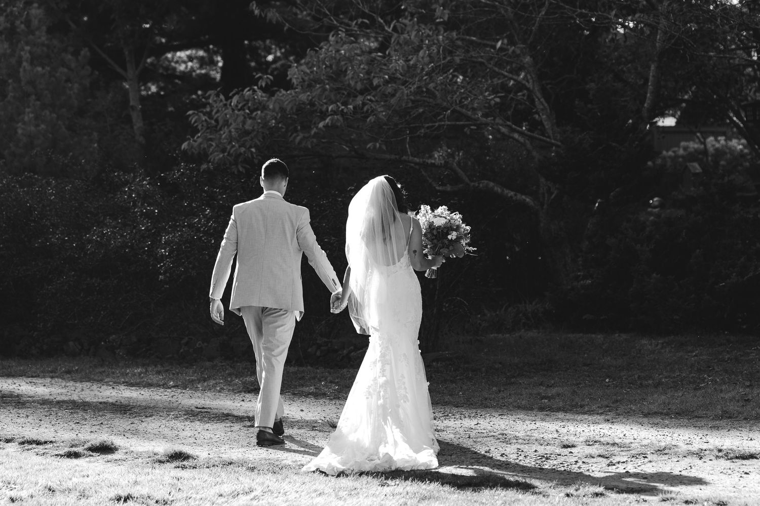Black and white photo of bride and groom walking hand in hand through a sunlit forest path.