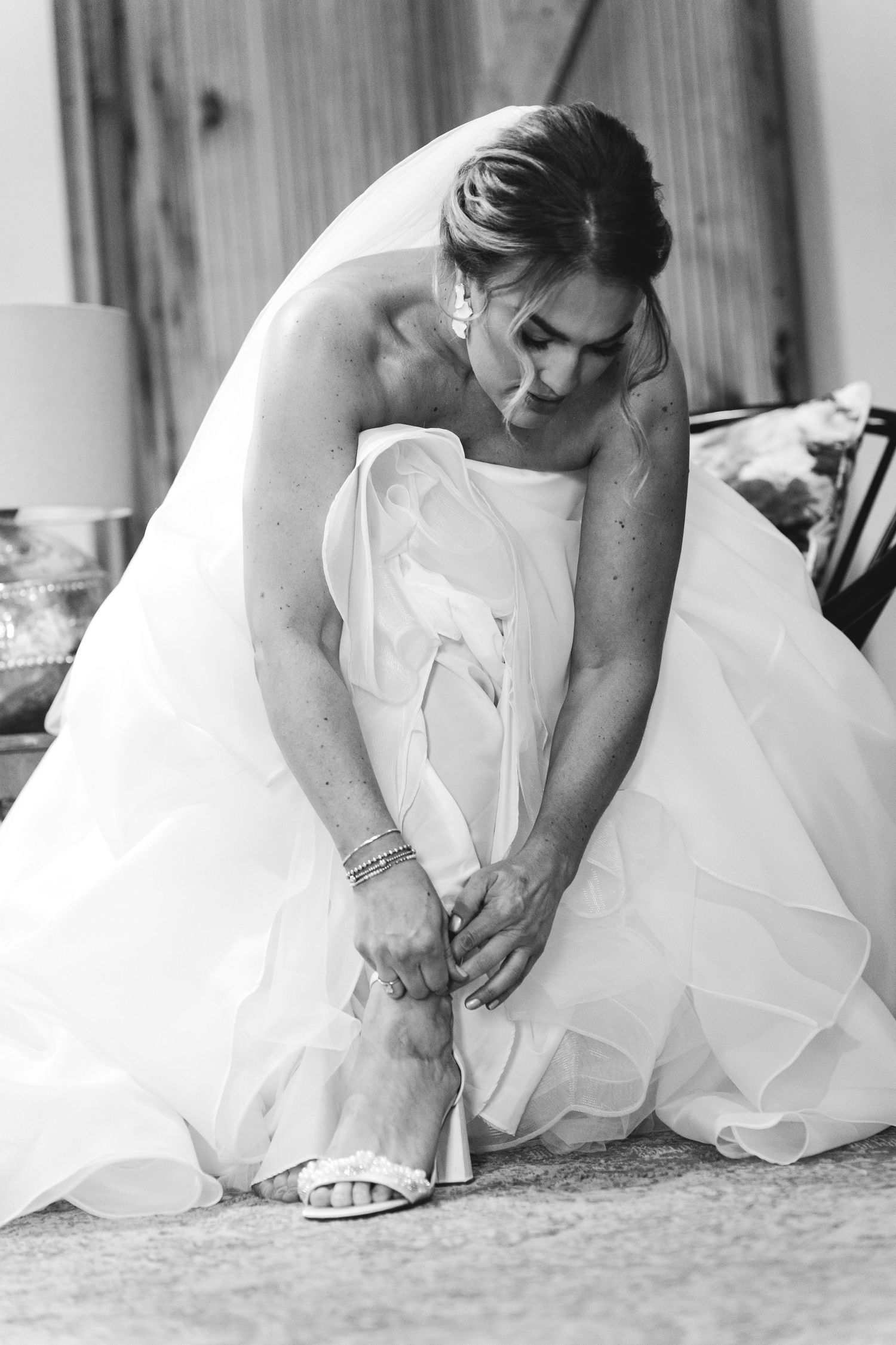 Black and white photo of a bride in a ball gown adjusting her heeled sandal on her wedding day.