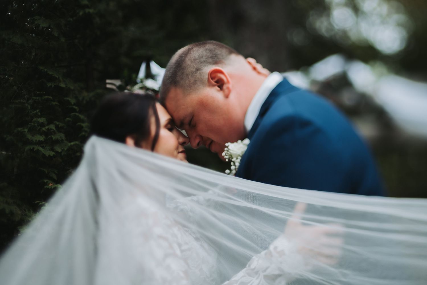 Romantic bride and groom sharing intimate kiss hidden beneath flowing white wedding veil outdoors.
