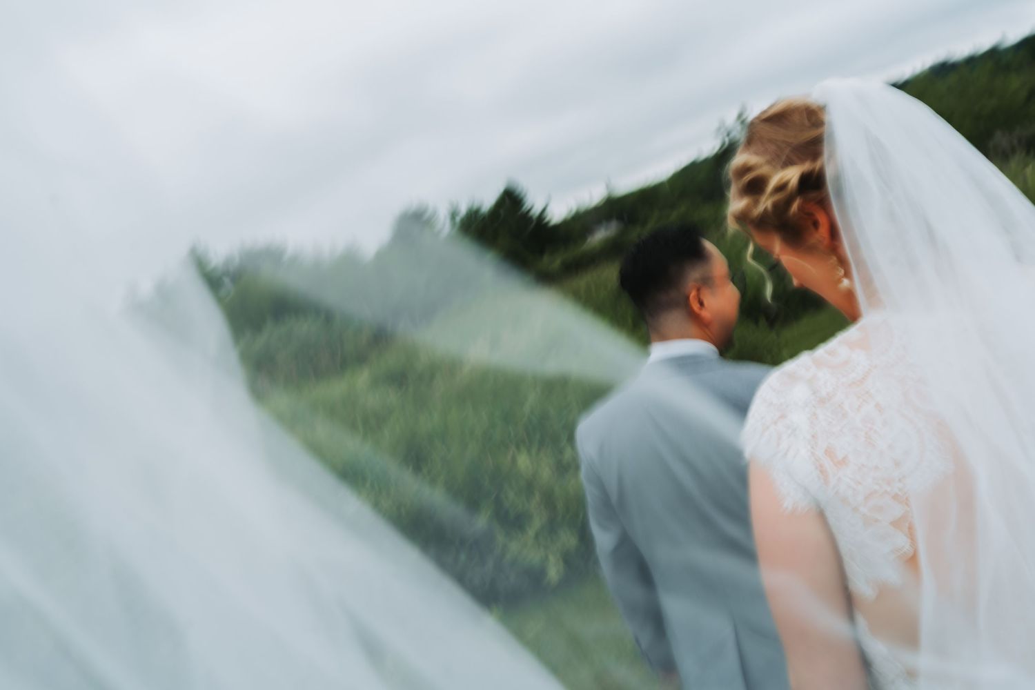 Bride and groom share a romantic outdoor moment as a flowing wedding veil billows in the wind.