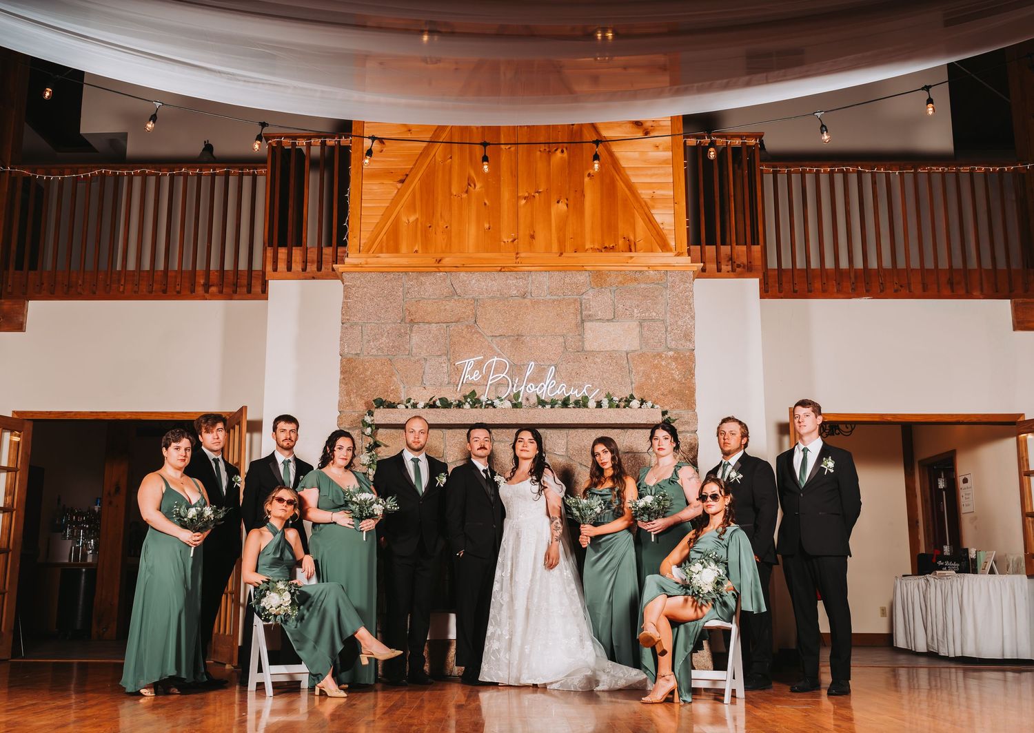 Wedding party poses in front of stone fireplace in elegant venue with green bridesmaid dresses.