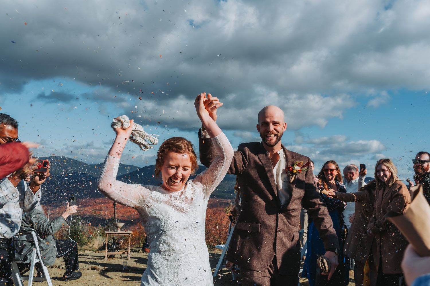 A joyful bride raises her hands as confetti falls during an outdoor wedding ceremony under a cloudy sky.