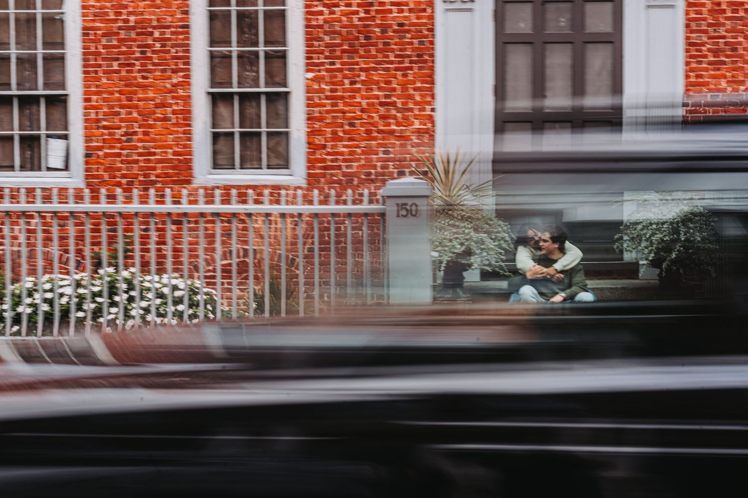 A couple sits on steps outside a red brick building, captured through motion blur of passing traffic.