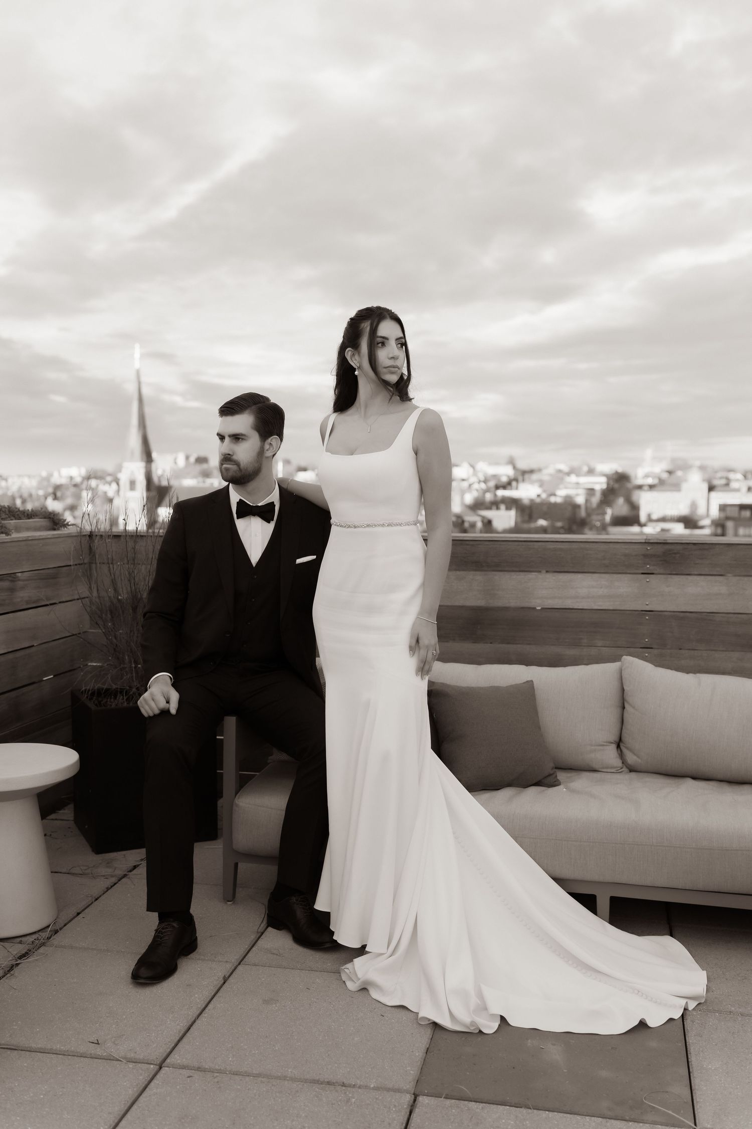 Elegant bride and groom posing on a rooftop terrace with city skyline backdrop in black and white wedding portrait.