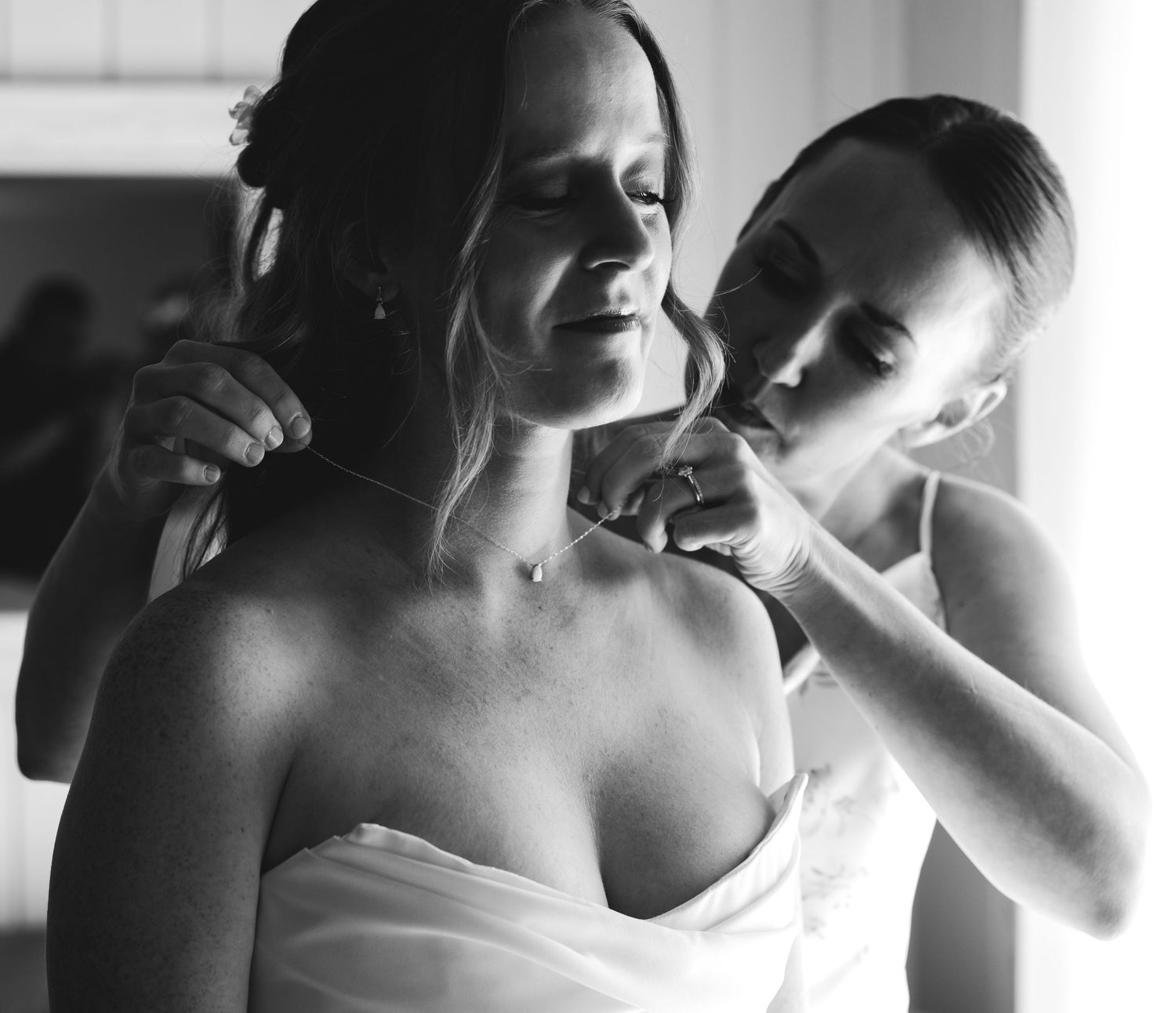 Black and white photo of a bridesmaid helping the bride with her necklace before the wedding ceremony.