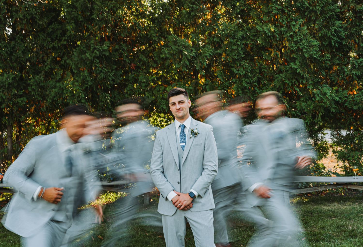 Groom stands still in a light gray suit while groomsmen blur past him in a dynamic wedding photo.