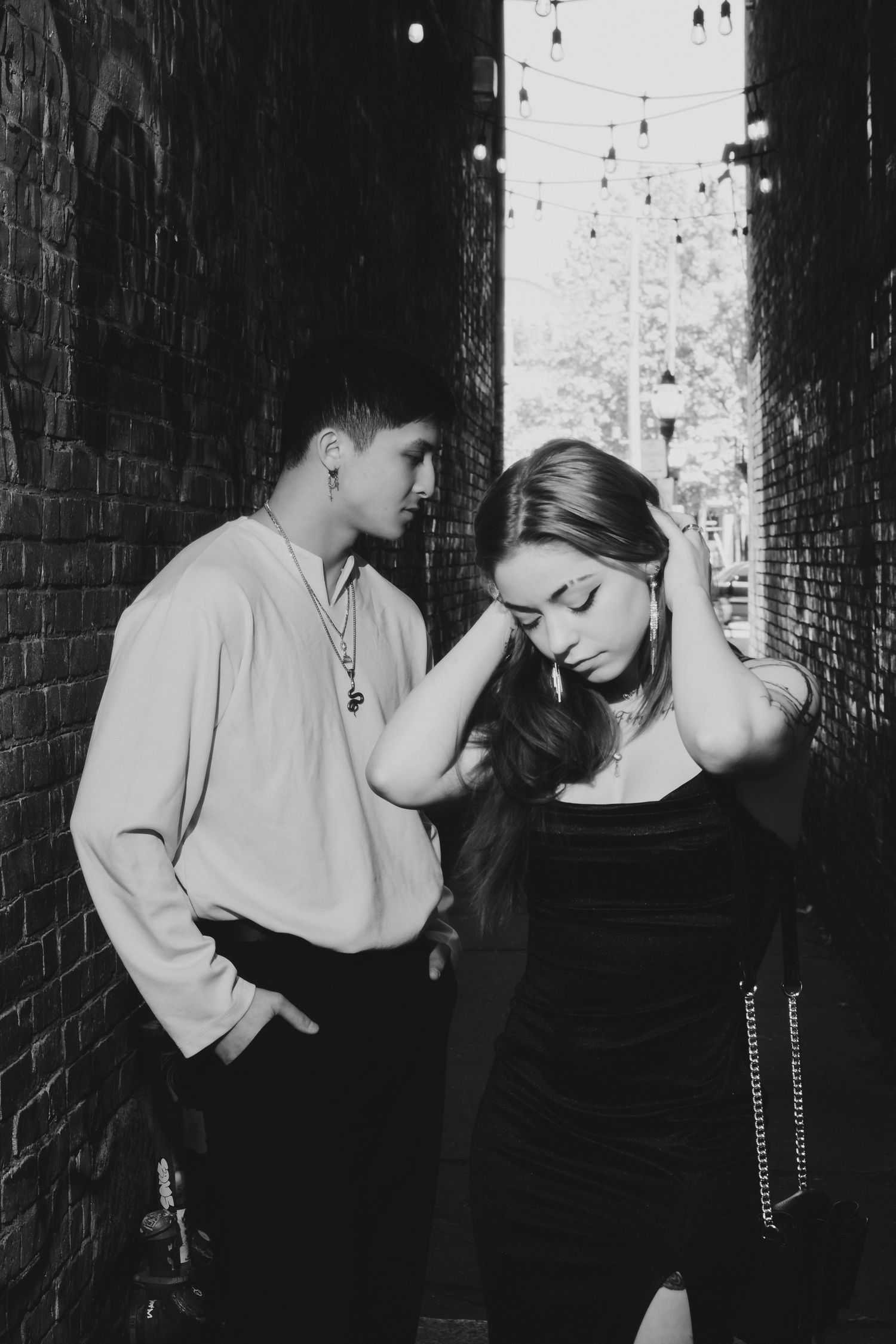 Black and white photo of a couple posing in a brick alley, the woman in a black dress with her hand in her hair.