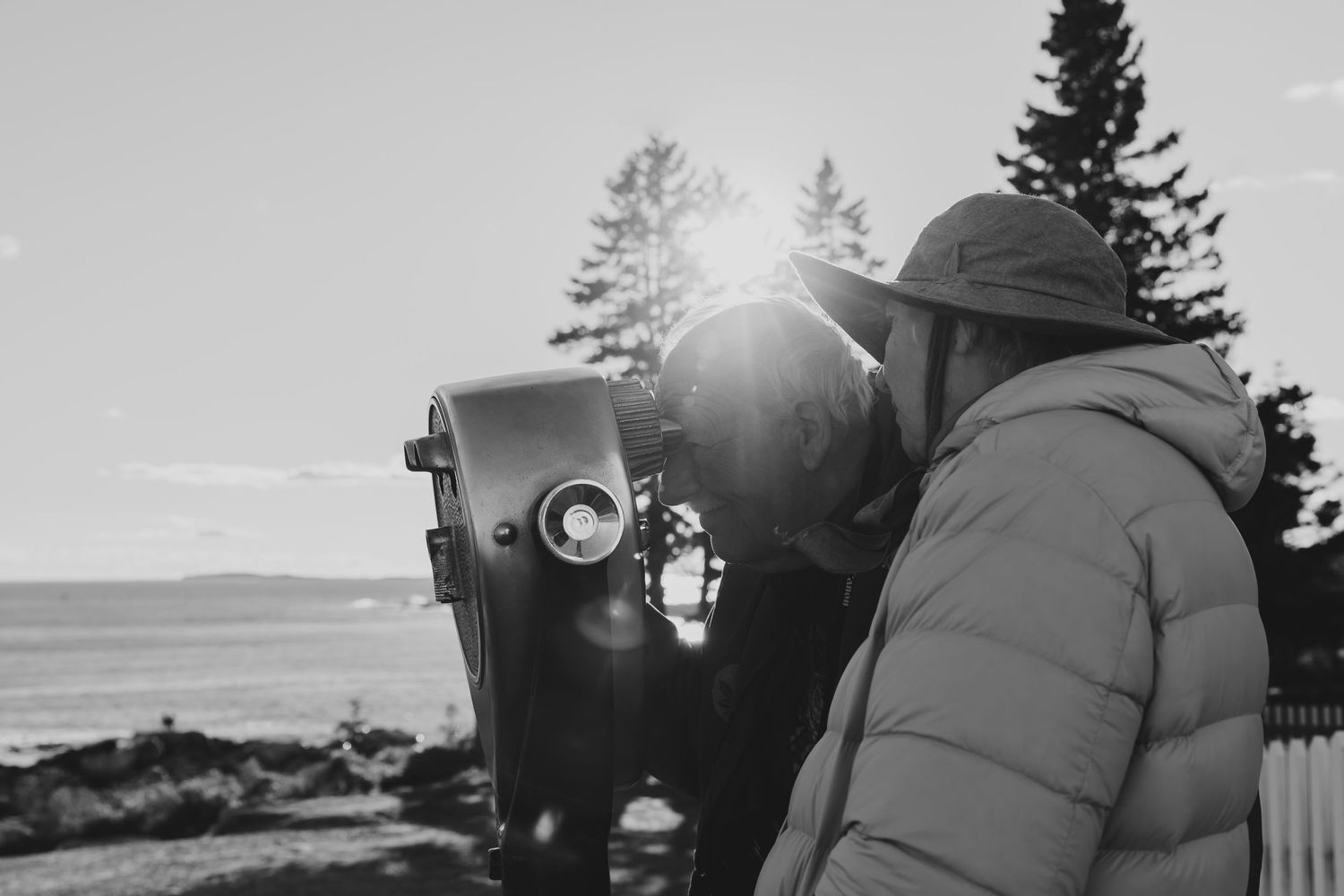 Person in puffer jacket using a coin-operated viewfinder binocular overlooking a scenic coastal landscape in black and white.