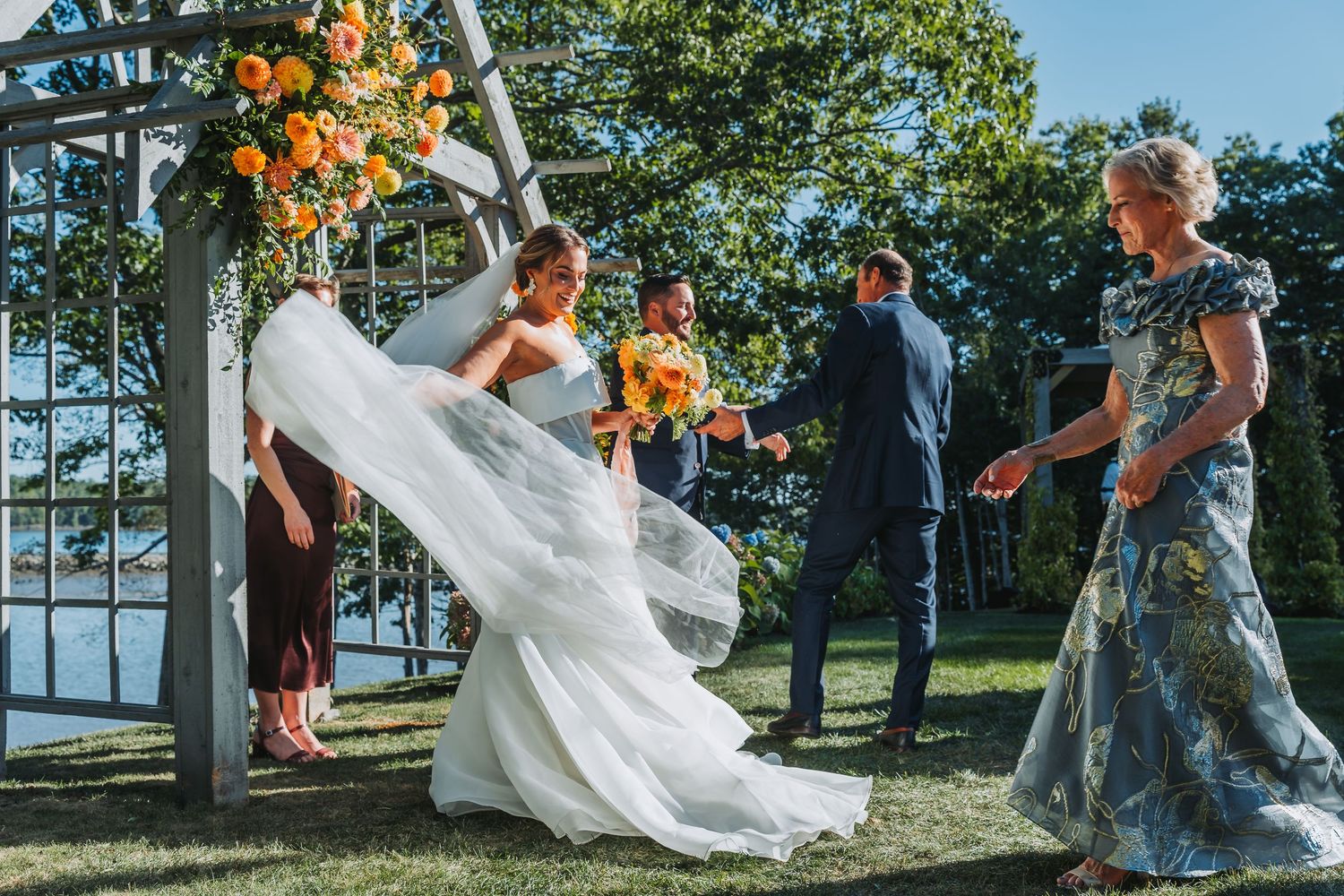 Bride in white gown with flowing veil at outdoor wedding ceremony, attendants holding orange bouquets near floral arch.