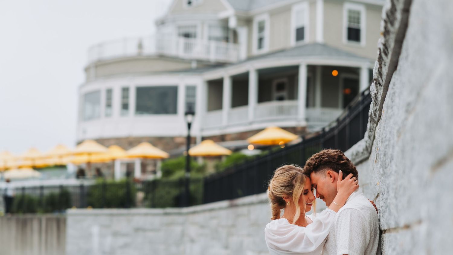 Couple shares an intimate embrace against a stone wall with a charming coastal wedding venue in the background.
