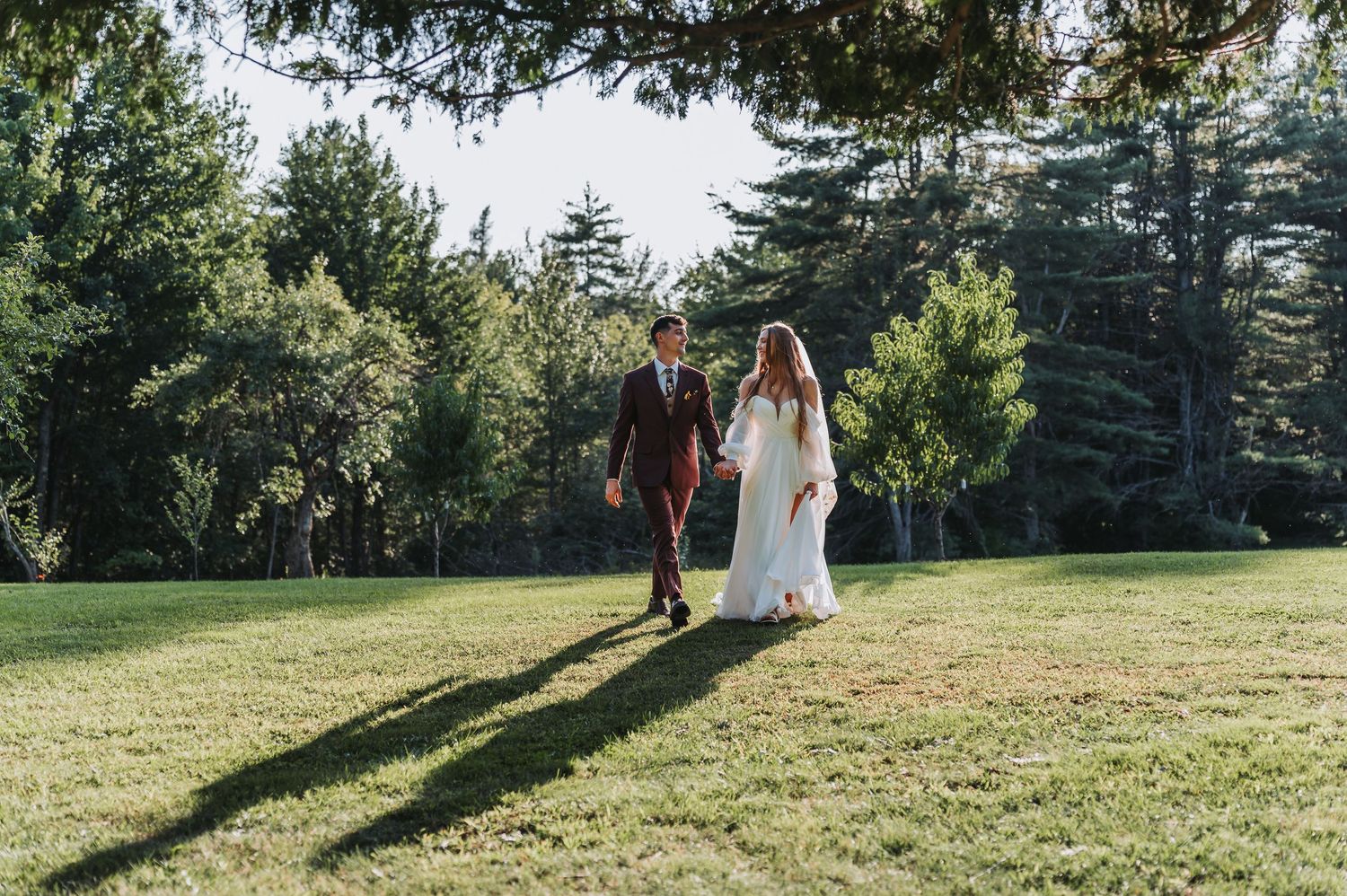 Bride and groom walking hand in hand across sunlit green lawn surrounded by tall pine trees.