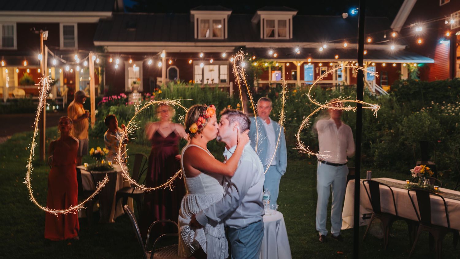 Newlyweds kissing under sparkler wedding signs at outdoor evening reception.