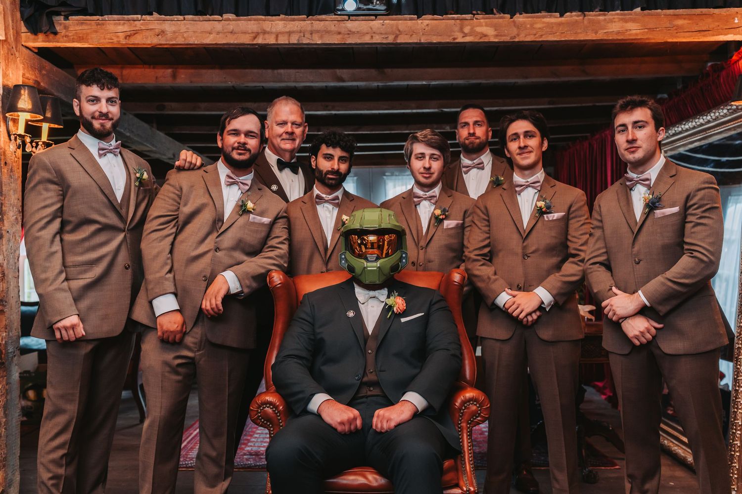 Groom seated in chair wearing helmet surrounded by groomsmen in brown suits with bow ties at rustic wedding venue.
