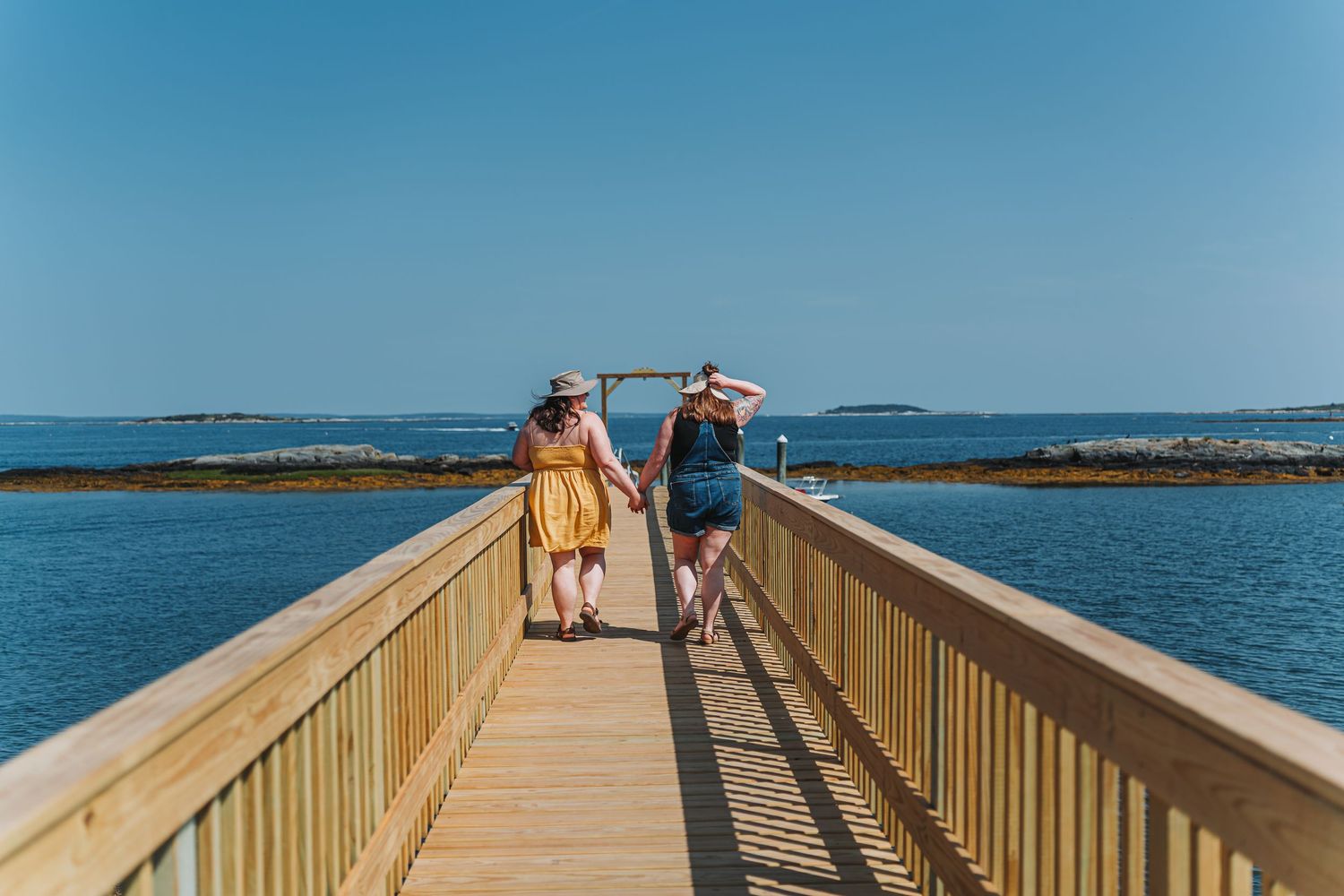 Two women walk along a wooden boardwalk overlooking a calm blue ocean on a sunny day.