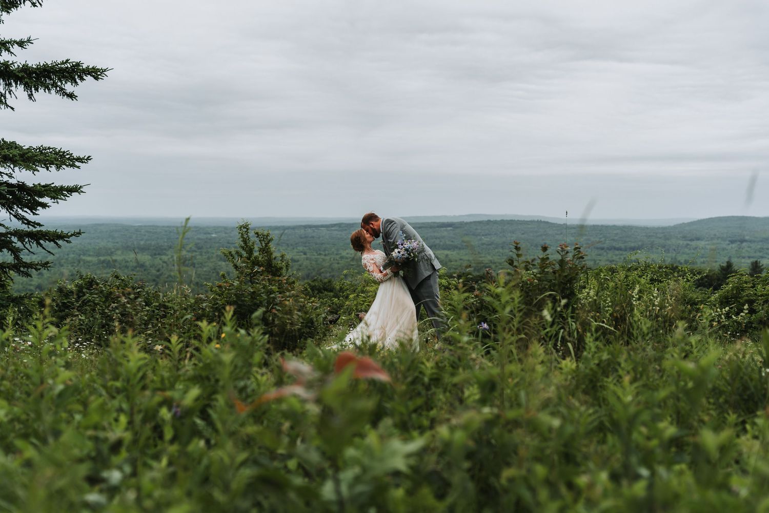 A couple shares a romantic kiss on a hilltop surrounded by lush green trees under an overcast sky.