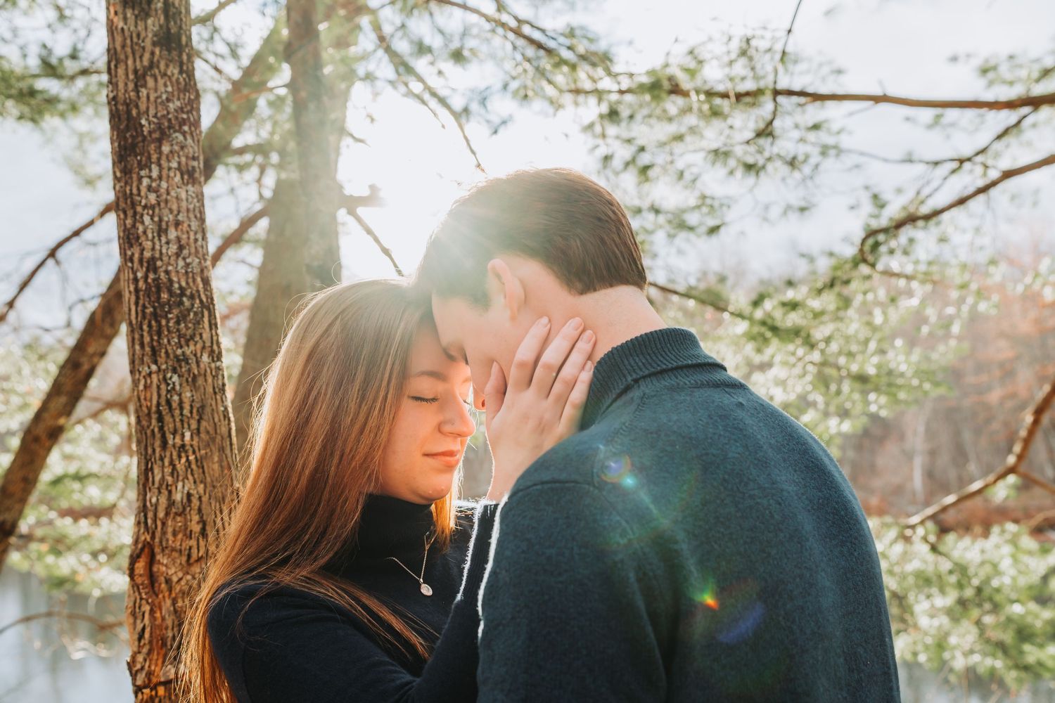 Romantic couple sharing a tender moment in sunlit forest, touching foreheads among pine trees.