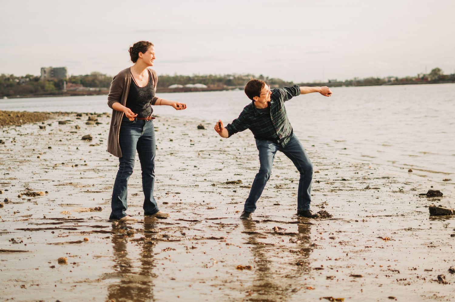 Couple playfully skipping stones on a muddy beach shoreline at dusk, laughing and enjoying a fun outdoor moment.