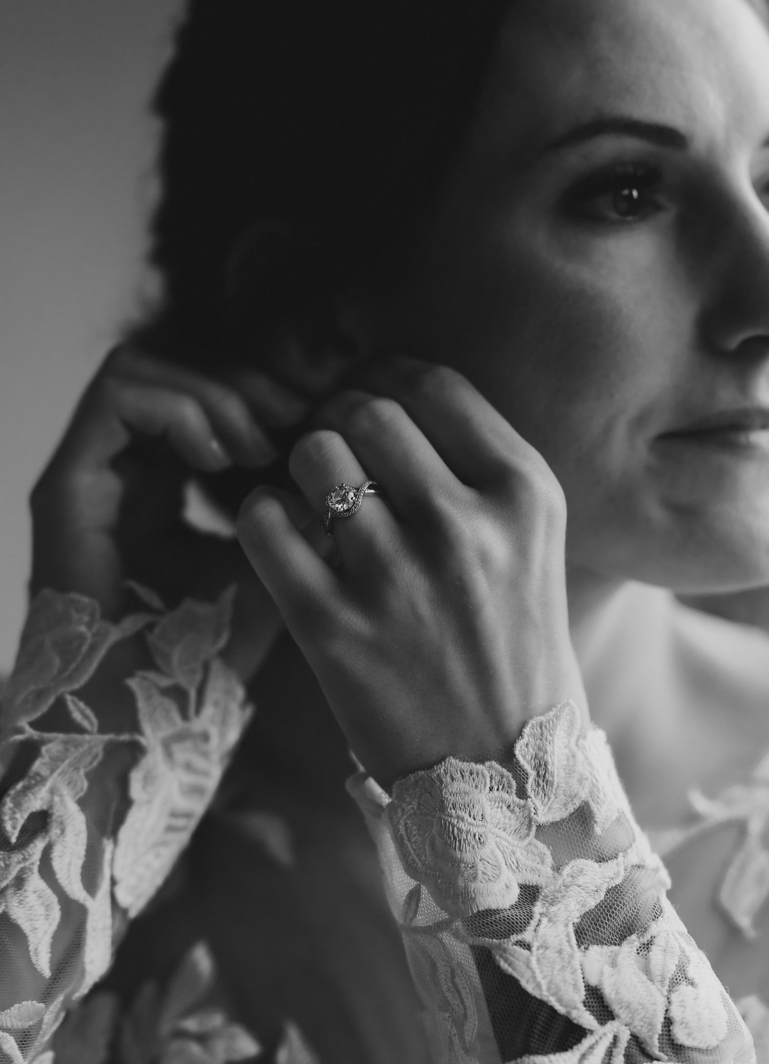 Black and white close-up of a bride adjusting her earring, wearing a lace gown and elegant ring.