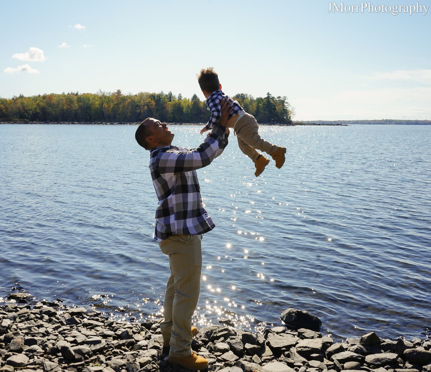 Father joyfully lifting baby in the air by a rocky lakeside on a sunny day.