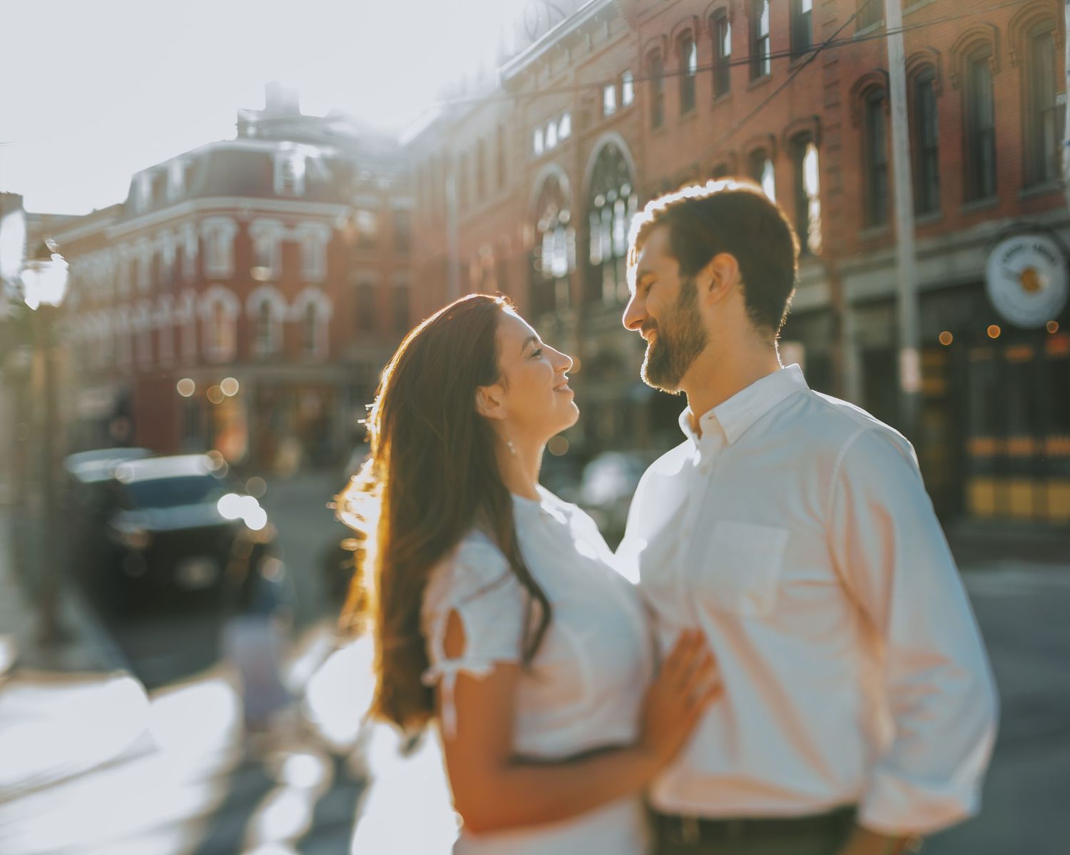Couple embracing and smiling at each other on a sunny urban street with a red brick building backdrop.