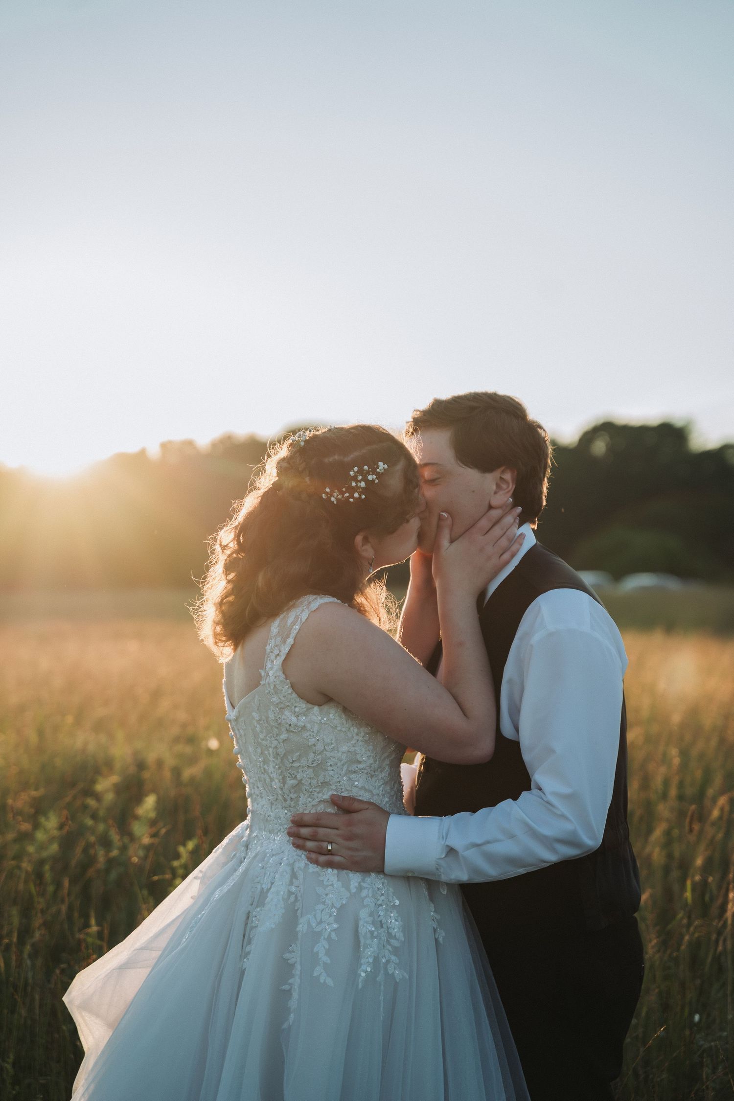 A bride and groom share a romantic kiss in a golden sunlit field during their outdoor wedding ceremony.
