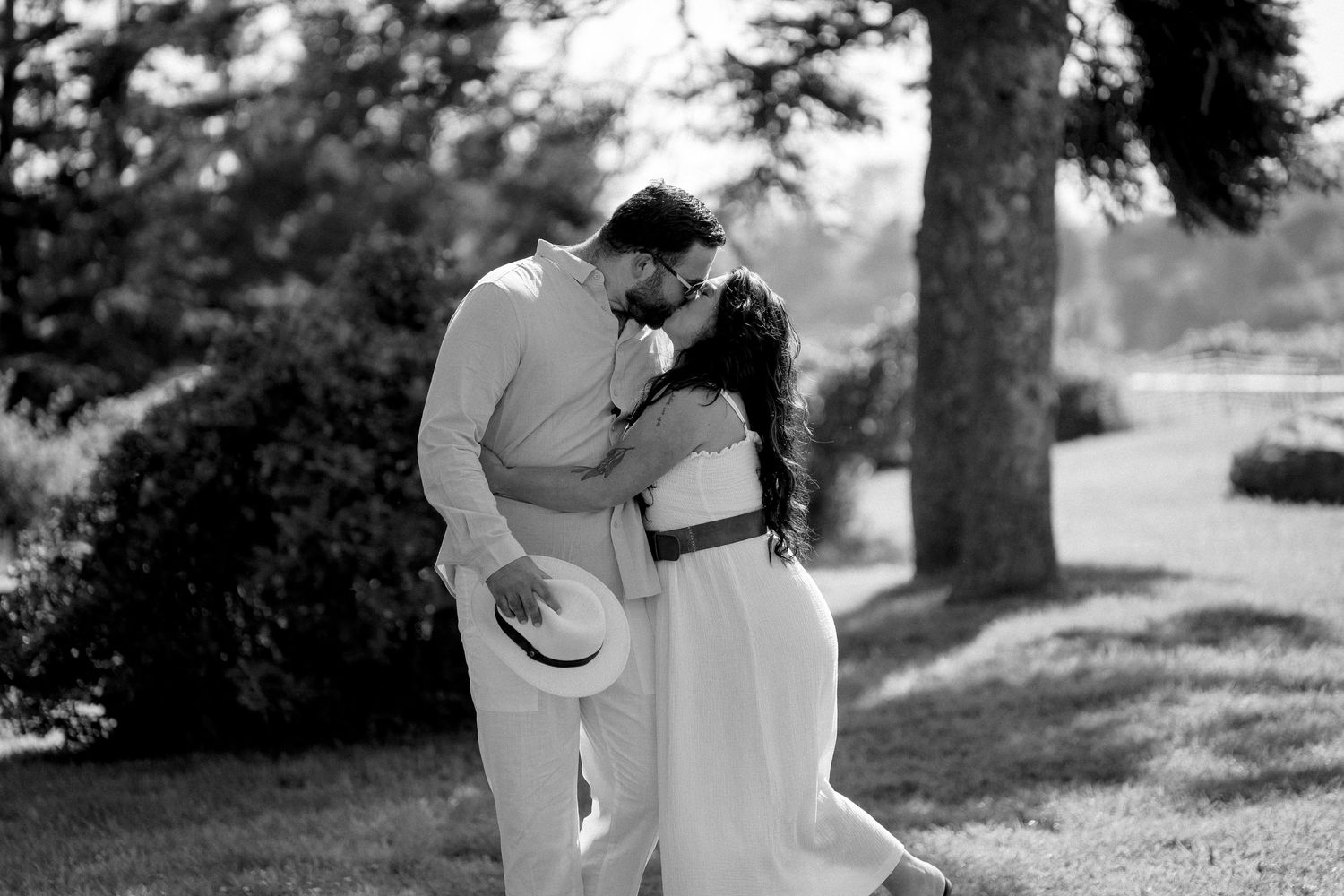 Black and white photo of a couple kissing in a lush garden, the man holding a hat while embracing the woman.