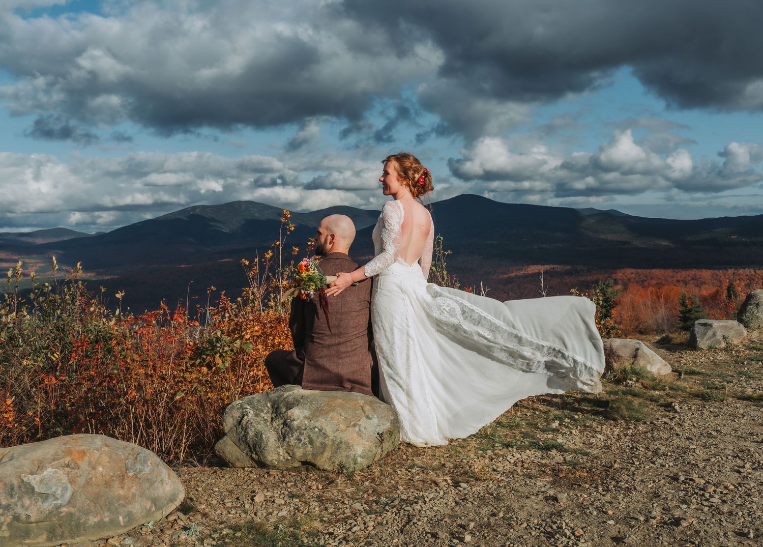 Bride in flowing lace gown with veil blowing in wind poses on mountain summit with stunning autumn foliage.