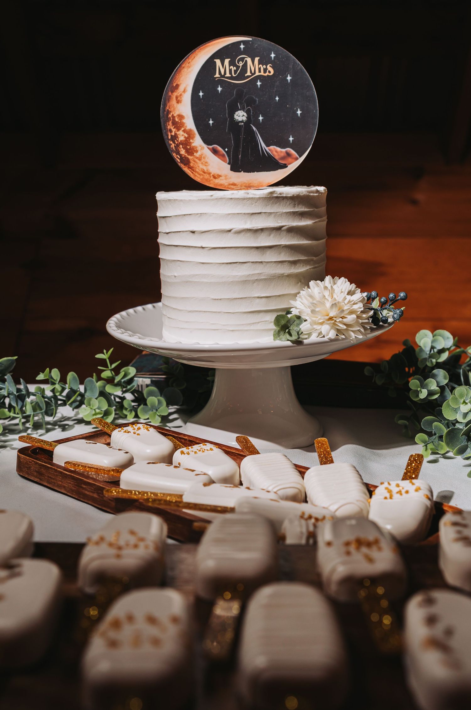 White single-tier wedding cake with Mr. & Mrs. moon topper on cake stand, surrounded by white frosted cookies.