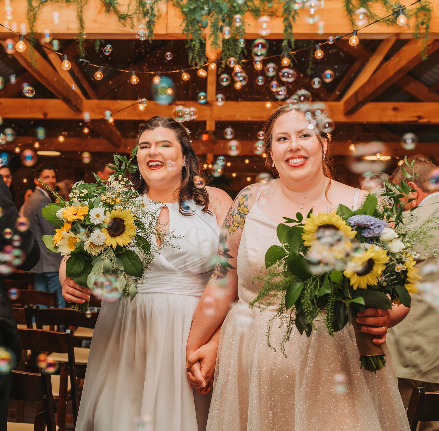 Two brides in white dresses holding sunflower bouquets walk down the aisle surrounded by floating bubbles.
