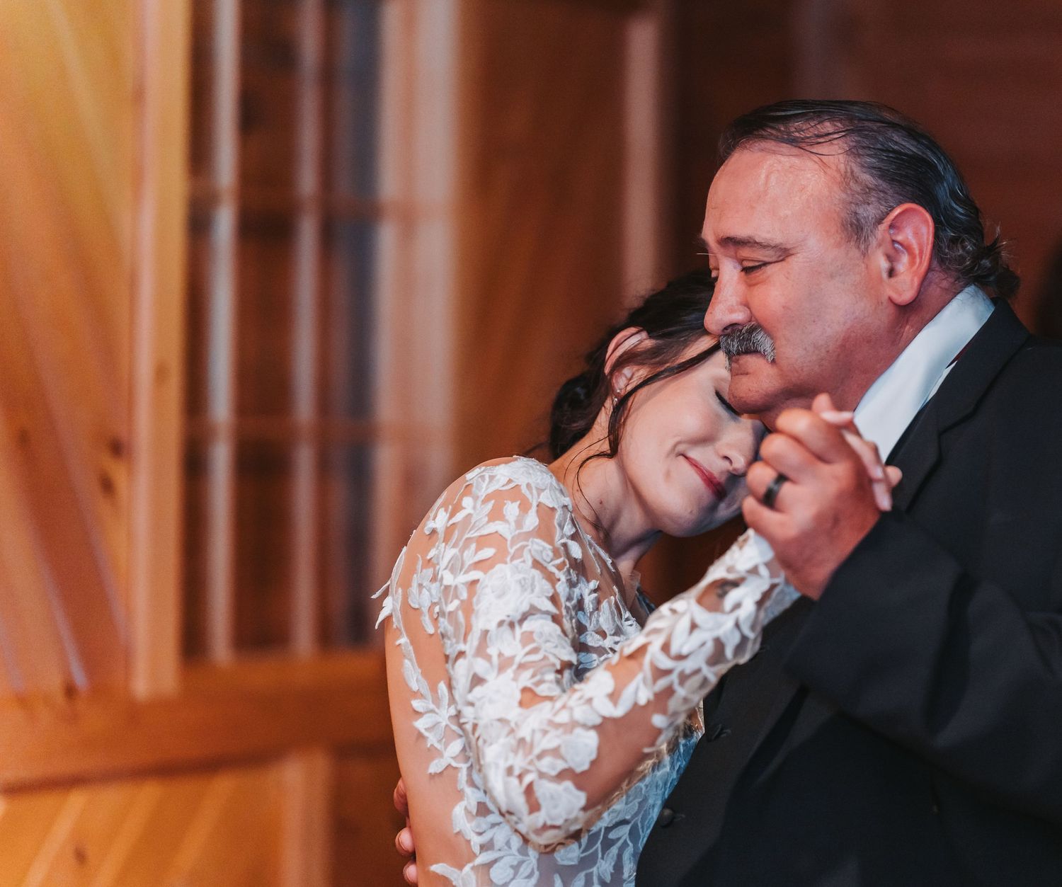 Emotional father-daughter dance with bride in intricate lace gown embracing during indoor wedding reception.