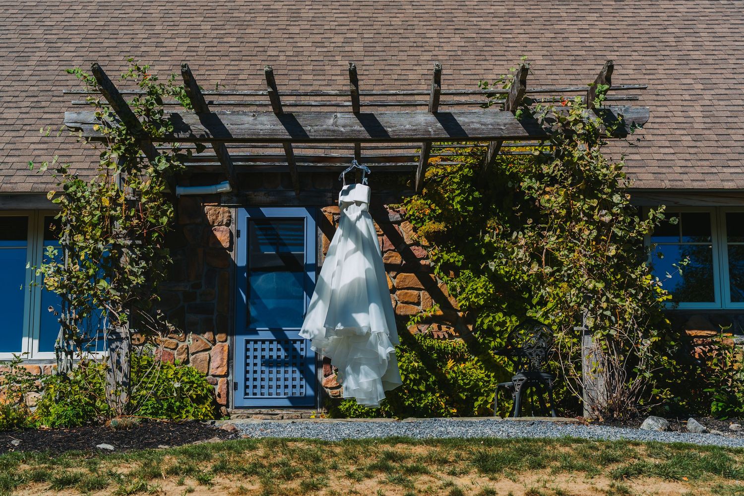 White wedding dress hanging under a wooden pergola on a brick building with a blue door surrounded by lush greenery.