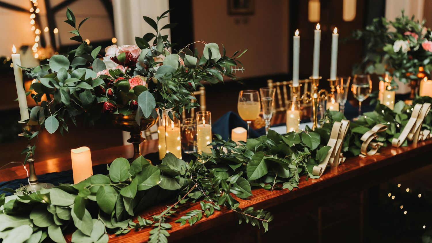 Elegant wedding table with greenery garland, white candles, gold accents, and floral centerpieces in a dimly lit venue.