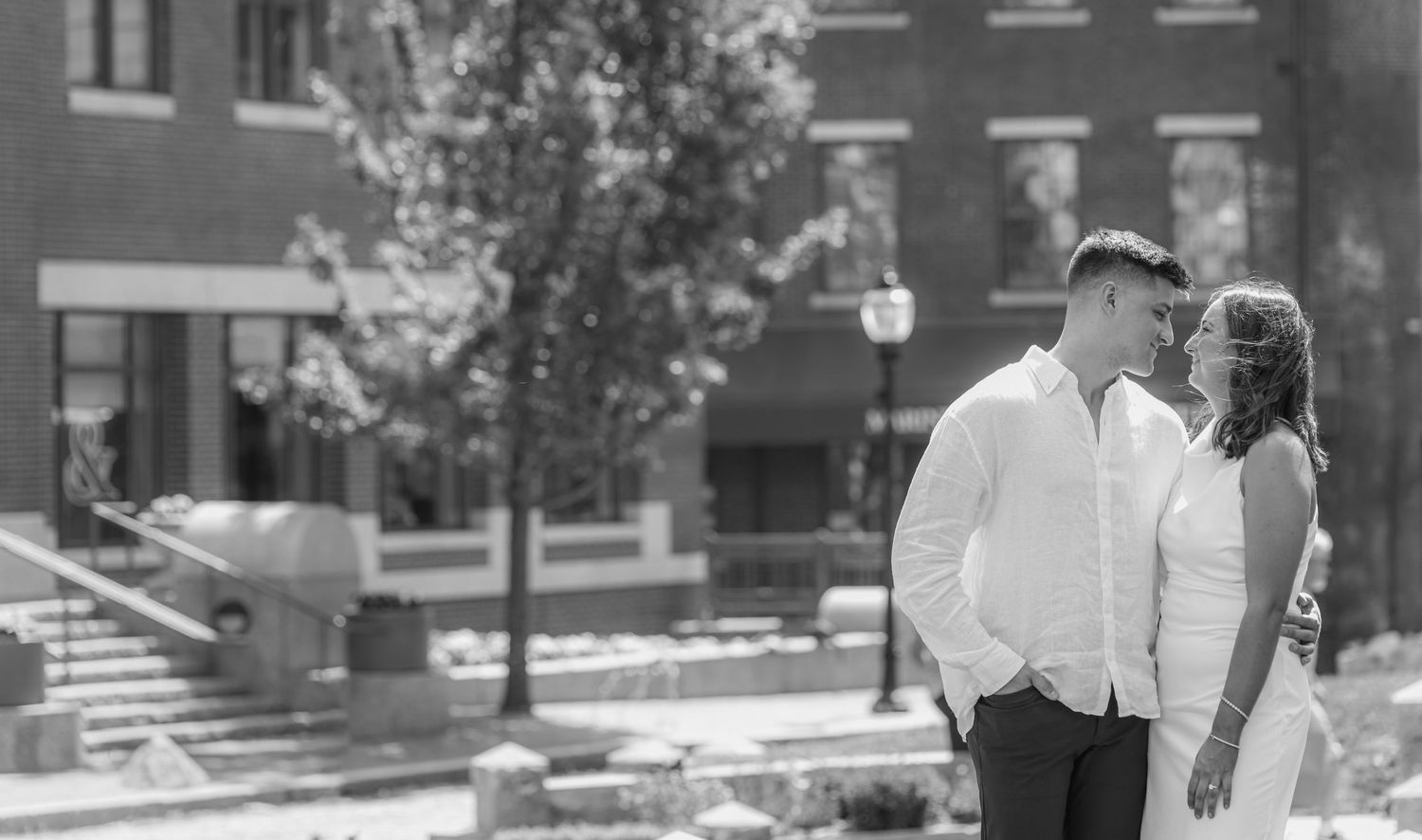 Black and white photo of a couple sharing a romantic moment outdoors near a brick building on a sunny day.