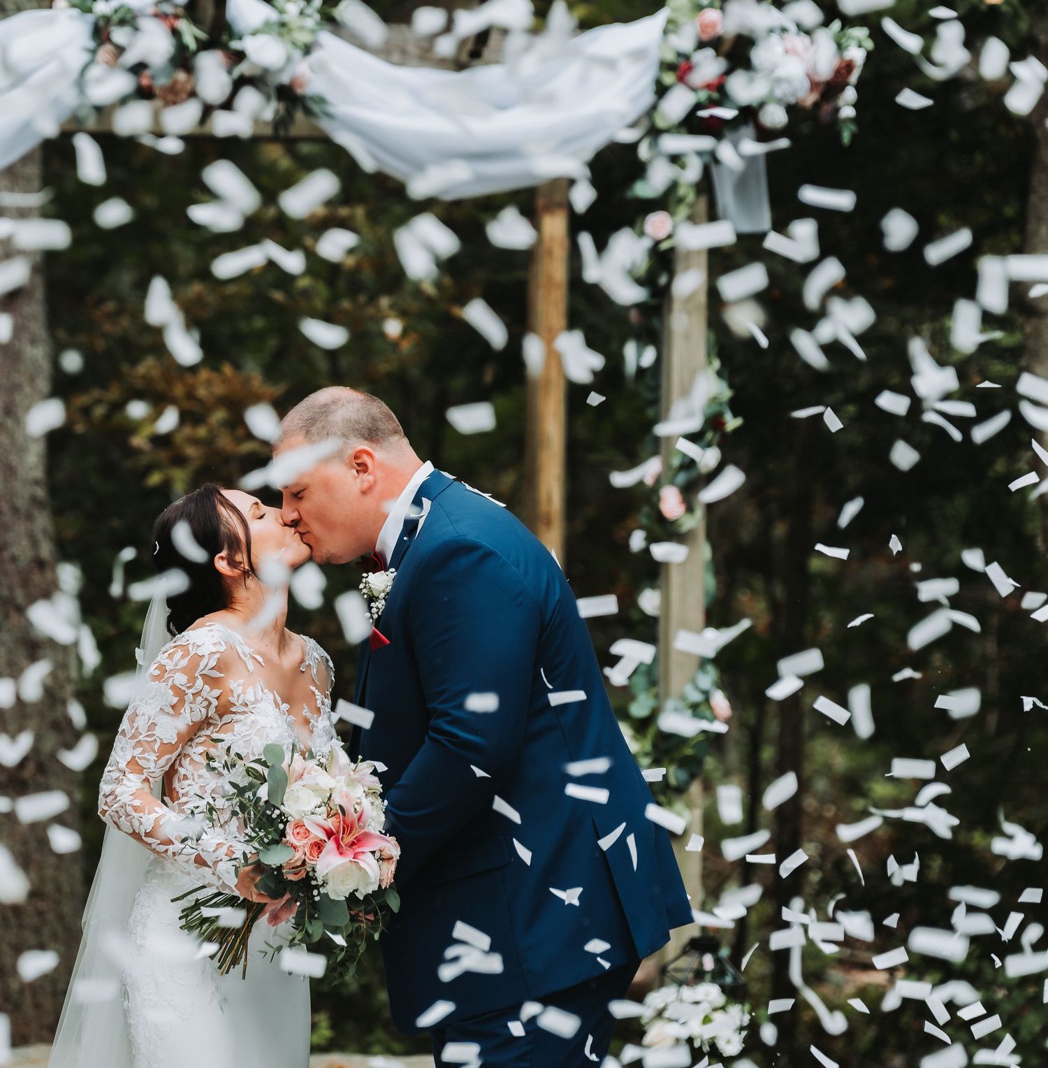 A bride and groom share a first kiss as white confetti fills the air during their outdoor wedding ceremony.