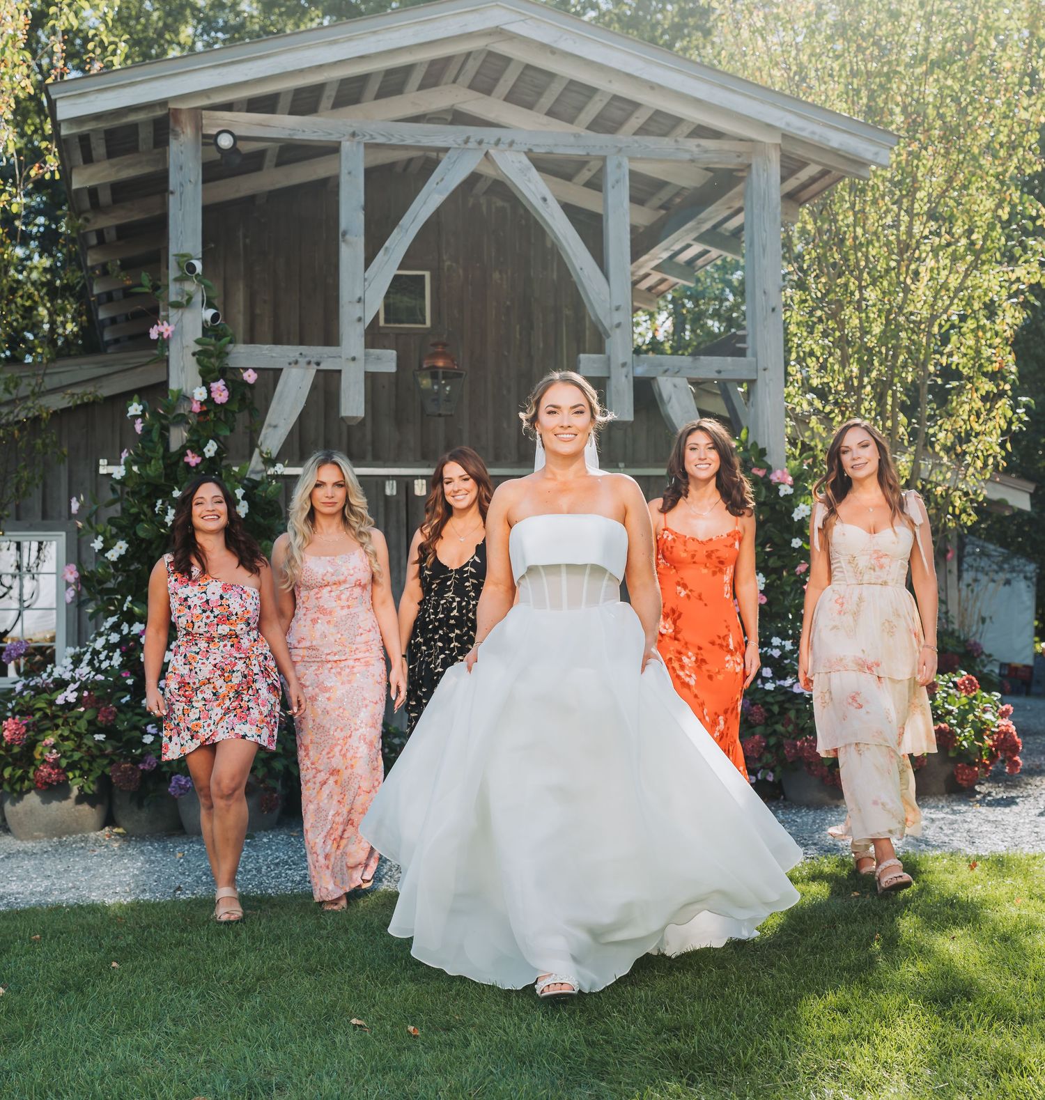 Bride in white gown walks with bridesmaids in colorful dresses outside rustic barn surrounded by flowers.