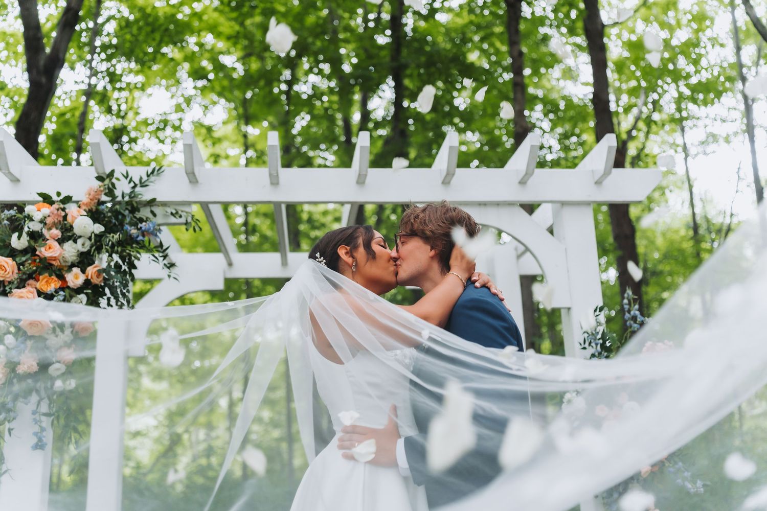 Bride and groom share first kiss under white pergola with veil billowing in the wind.